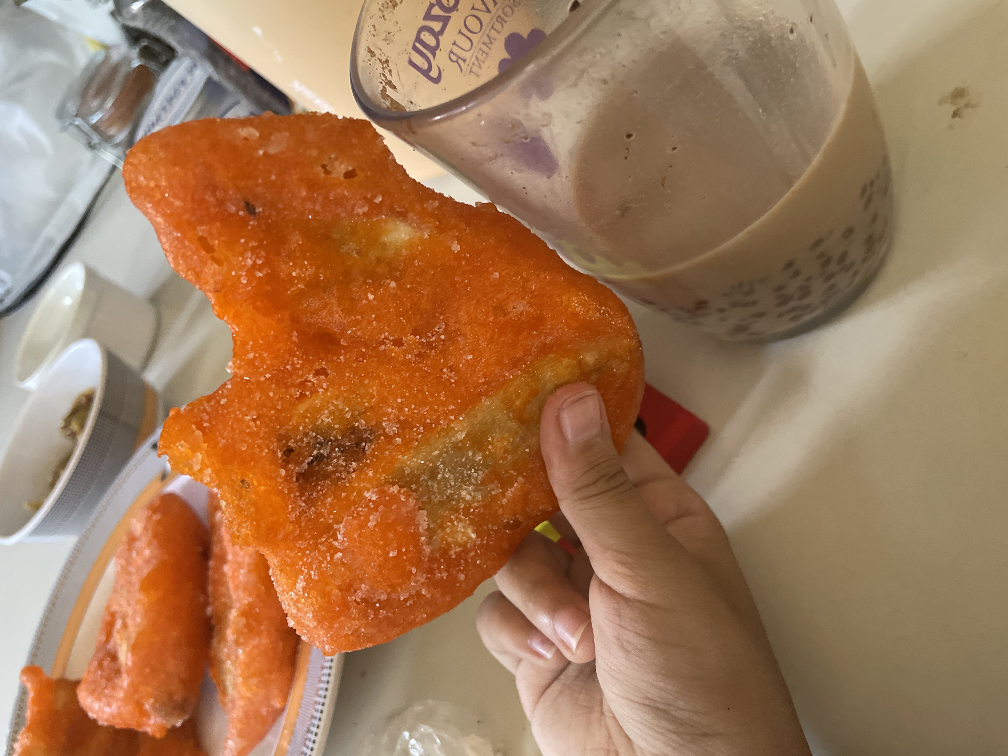 A person holds a piece of camote cue (candied sweet potato) in their hand, with more camote cue on a plate to the left. A glass of milk tea with tapioca pearls sits to the right of the hand. The setting appears to be indoors, likely a home, in Davao City, Philippines, judging from the food commonly found there. Other items are visible in the background, including a bowl and a food container.