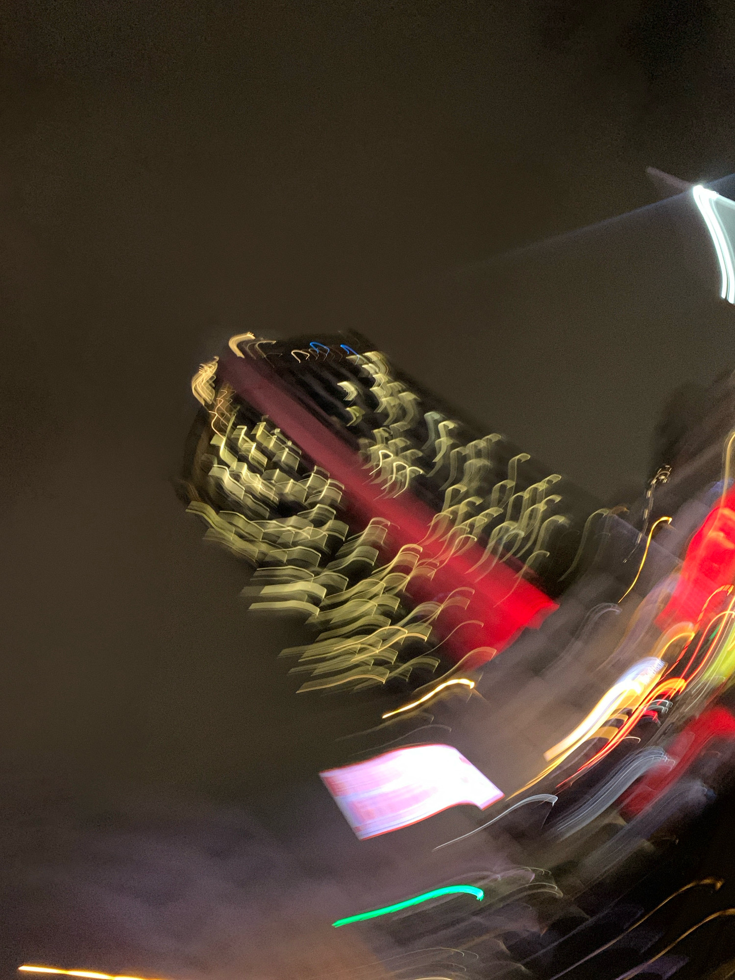 This is a long-exposure photograph of a city at night, likely taken from a moving vehicle or with camera movement. The image captures the blurred streaks of light from buildings, traffic, and signage in the Beyoğlu district of Istanbul, Turkey. The dominant colors are the warm yellows and golds from building lights, contrasted with vibrant reds and greens from signs and vehicle lights. The dark, nebulous sky suggests a cloudy or hazy night. The overall impression is one of dynamism and the abstract beauty of urban nightlife.