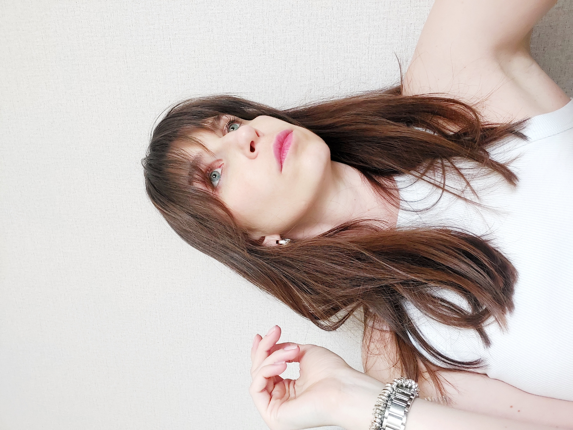 A woman with shoulder-length brown hair and bangs lies against a white textured surface. She wears a white tank top and a silver bracelet. Her face is tilted upwards, revealing light blue eyes and pink lips. She is wearing stud earrings. The image was taken at an angle, not allowing for confirmation of a specific location in the city of Chaikovsky, Russia.