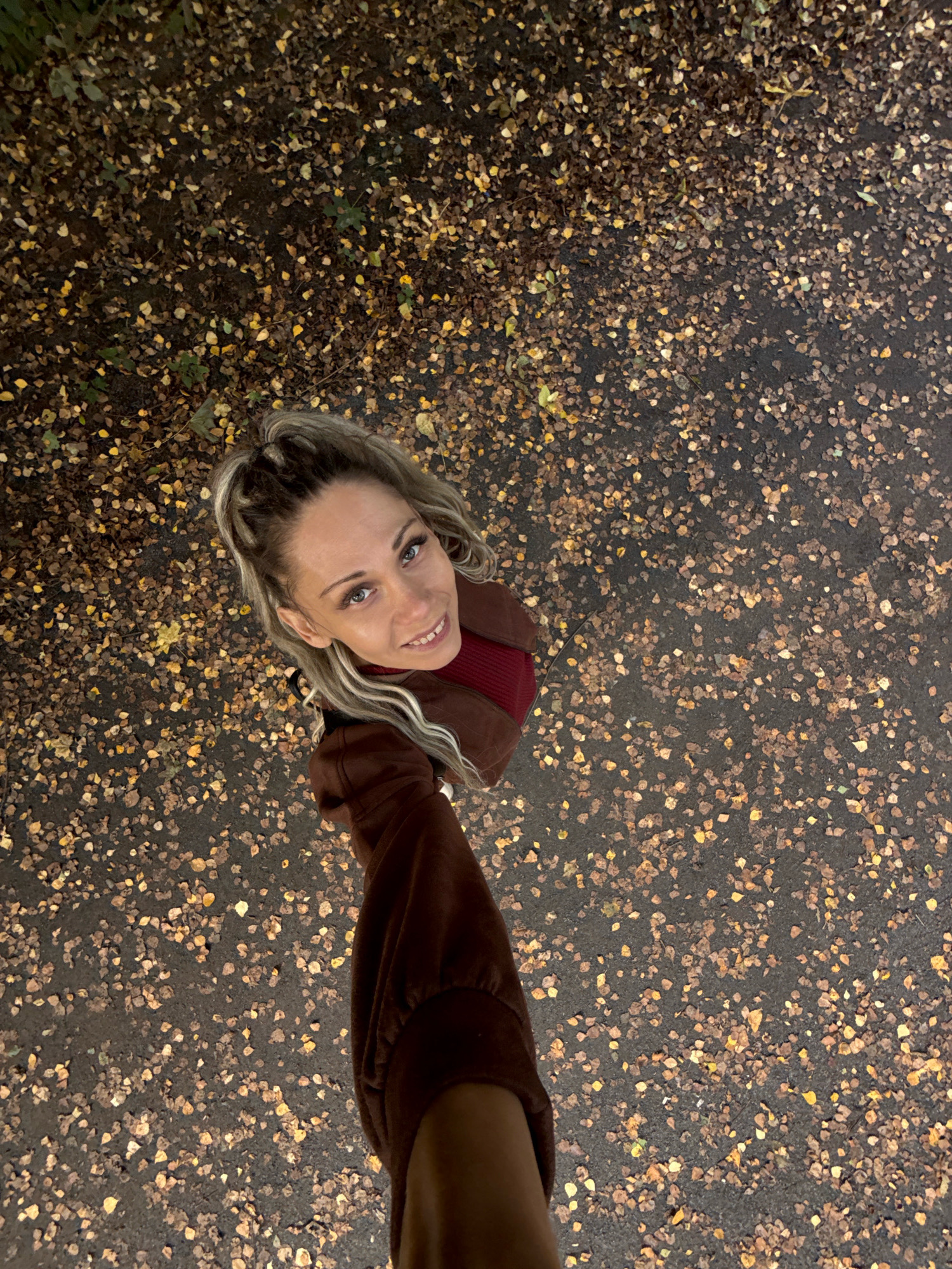 A woman with blonde hair tied in a high ponytail smiles at the camera, looking up from a slightly high angle. She is wearing a reddish-brown jacket over a dark red turtleneck. The background is a dark asphalt path covered with fallen autumn leaves in shades of brown and yellow. The scene appears to be outdoors in a park or along a tree-lined path, likely in Rusanovo, Russia, given the typical deciduous foliage and ground cover associated with Russian autumns.