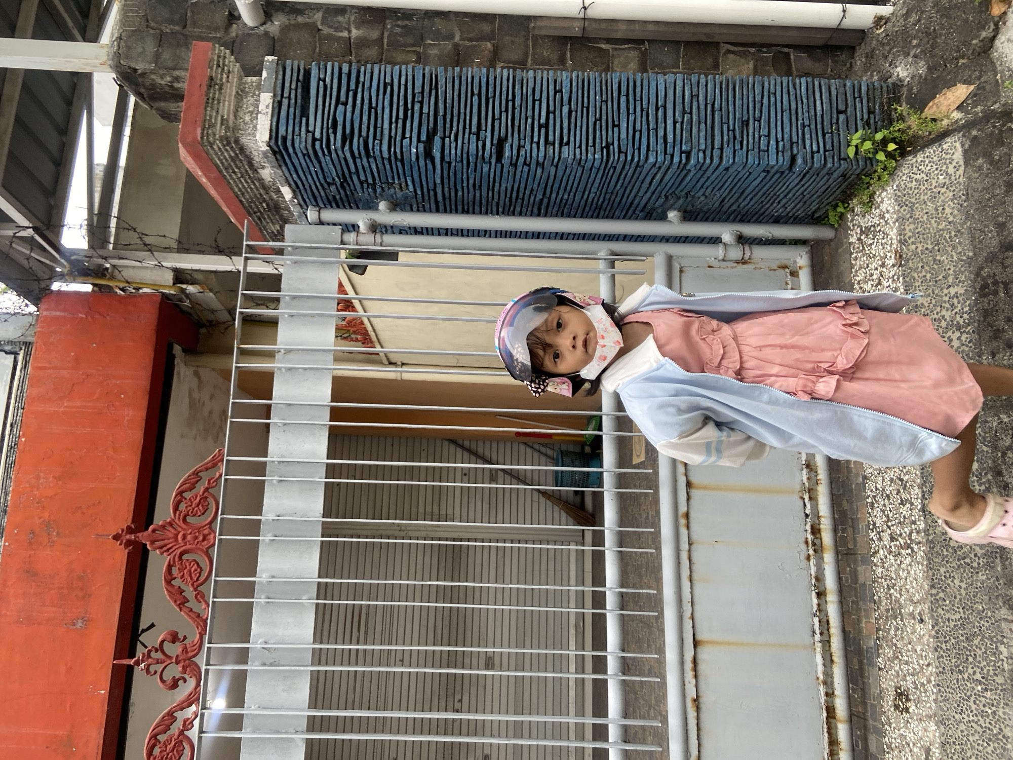 A girl stands in front of a gray metal gate with horizontal bars in Surabaya, Indonesia. She wears a light pink dress, a light blue jacket, a helmet with a visor, and a face mask. The gate is situated between a red decorative wall on the left and a blue tiled structure on the right, with a paved walkway beyond. The background shows glimpses of other buildings and vegetation.