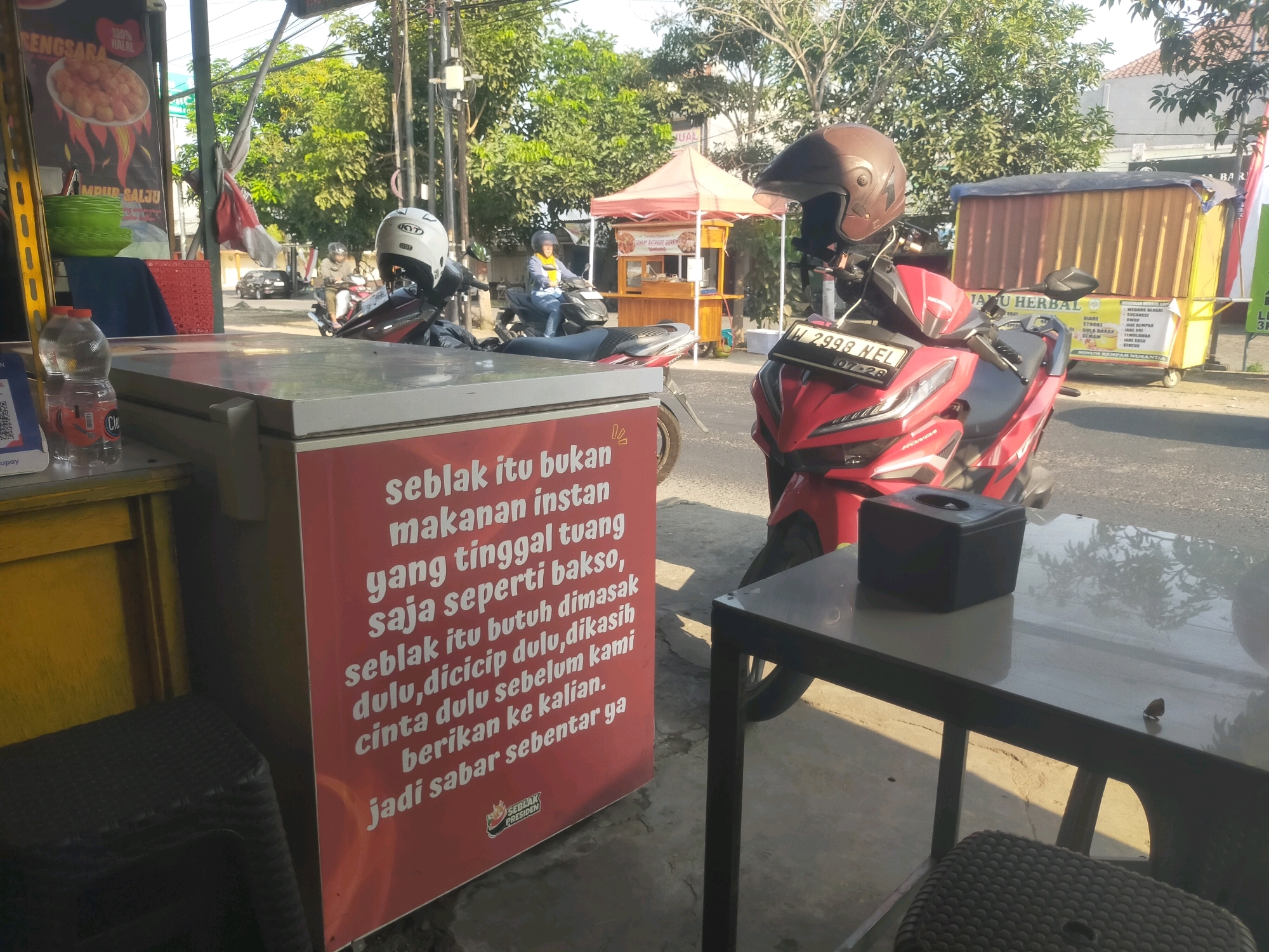 In Sidodadi, Indonesia, an outdoor scene depicts a roadside food stall with several motorcycles. A red scooter with the license plate H 2998 NEL is parked prominently in the foreground, its brown helmet resting on the handlebars. Another scooter, white with a black helmet, is parked behind it. Behind the motorcycles, a food cart with a red awning and wooden structure is visible, displaying food items. A yellow container on wheels is positioned further back, with signage indicating "JATU HERBAL". The food stall features a red sign with white text describing "Seblak" and its preparation. In the immediate foreground, a white cooler is partially visible, with a bottle of water and what appears to be a QR code attached. A black table with a tissue box sits in the right foreground. The environment is characterized by trees, a paved road, and buildings in the background, suggesting a daytime setting with sunlight casting shadows.