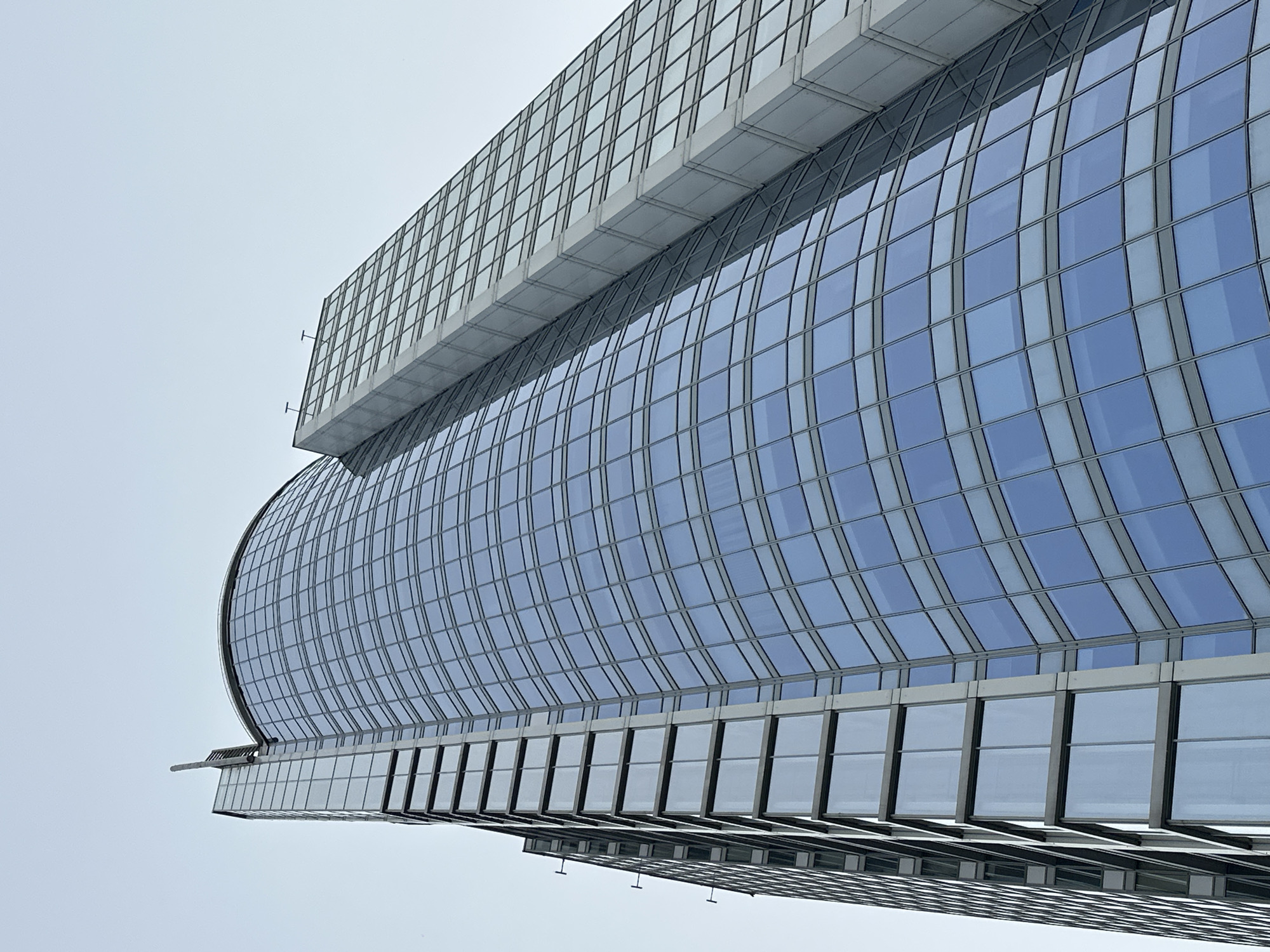The image presents a low-angle, upward-looking perspective of a modern skyscraper, identified as the Skyline Tower (Skajlajn) in Minsk, Belarus. The building's facade is predominantly composed of reflective, blue-tinted glass panels set within a grid of grey-silver metal framing. The left side of the structure features a prominent, continuous curved glass surface, with numerous rectangular window sections. At the very top left of this curved section, a slender, vertical architectural fin extends upwards. To the right, the building exhibits a more rectilinear or angular design, also clad in glass, but with distinct horizontal bands of grey material separating sections. The overall architectural style is contemporary, emphasizing clean lines and a sleek, transparent appearance. The background consists of a clear, light blue to white sky, providing a stark contrast to the dark blue reflections on the building's glass. No people, vehicles, or other surrounding urban elements are visible due to the tight, upward crop.