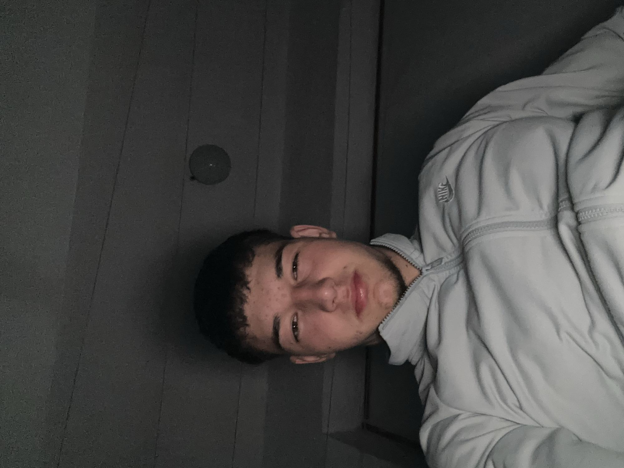 A portrait of a young man is captured in a dimly lit, indoor setting. He is wearing a light gray Nike jacket. The background shows a gray surface with vertical lines, potentially a wall or a door. There is a circular object mounted to the surface behind him. Location details in Zlatograd, Bulgaria are not discernible from the image.
