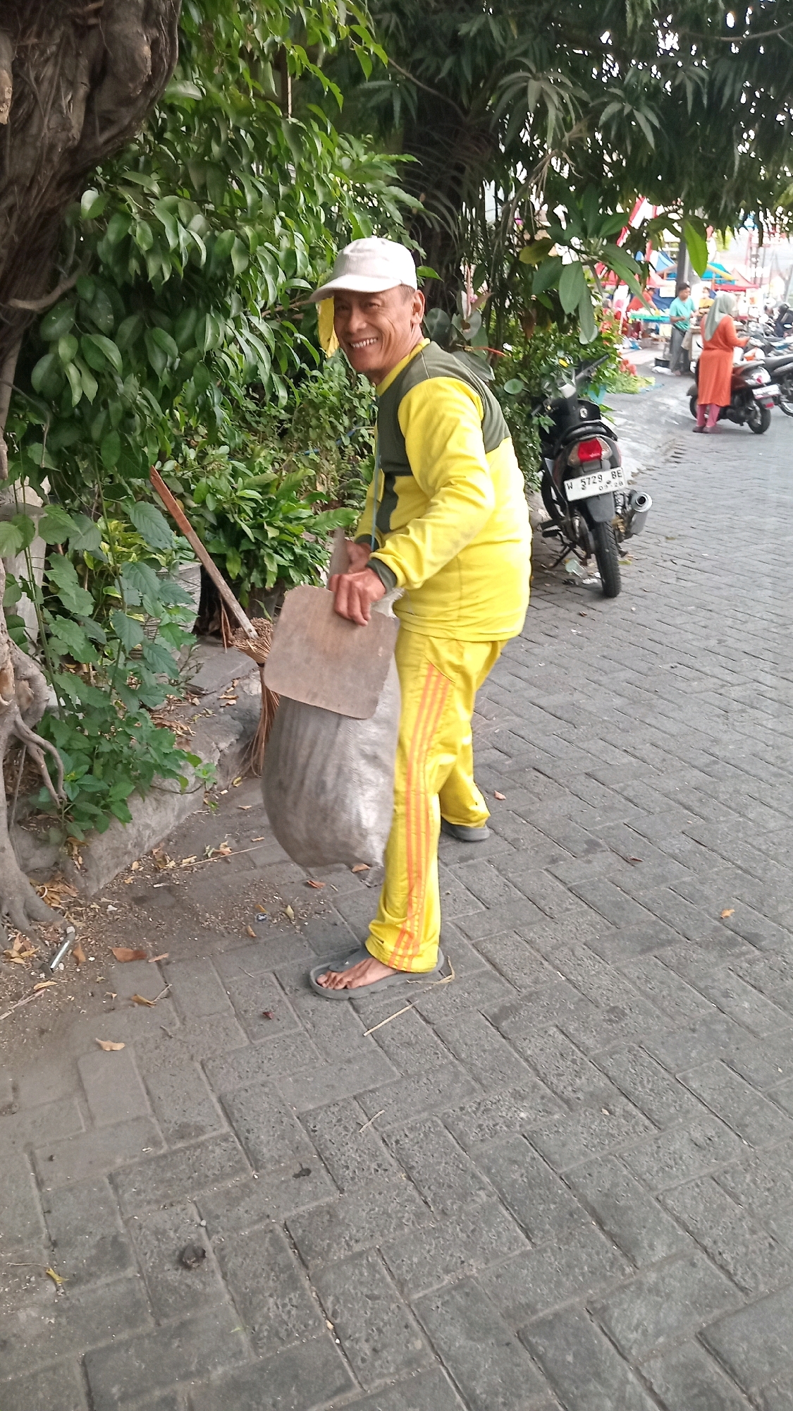 A man in a yellow uniform with orange stripes on his pants is holding a dustpan and a large gray garbage bag on a paved walkway. He is wearing a white cap and smiling at the camera. Behind him, there are trees with green leaves, a motorcycle, and two people standing in the background near what appears to be a market stall. The walkway is made of interlocking gray pavers.