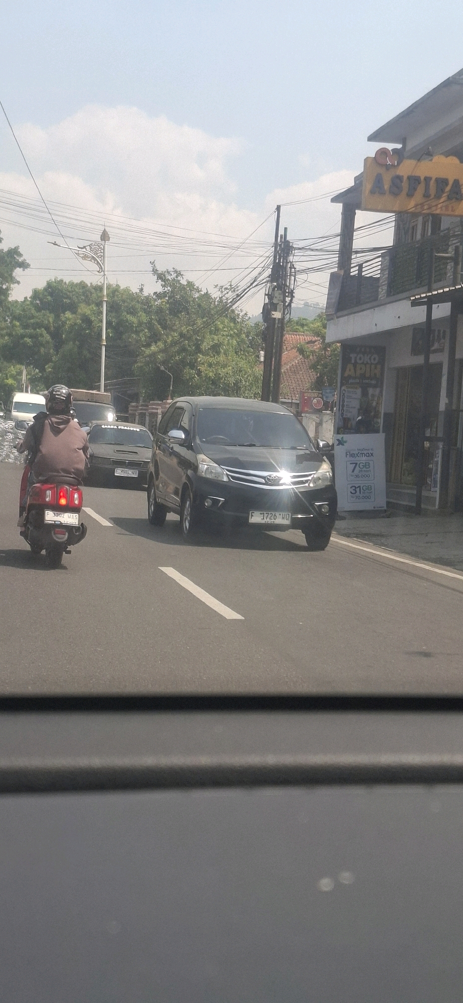 A black Toyota Avanza with license plate F 1726 HQ is driving on a street in Sukasari, Indonesia. The car is in the right lane, with a white vehicle ahead of it and a red scooter to its left. The scooter, with license plate F 3302 WAS, is carrying a person wearing a brown hoodie. The road is paved, with white lane markings. On the right side of the road, there is a building with a sign that reads "ASPIFA" in large yellow letters. Below the sign, there is a storefront with a sign that says "TOKO APIH". Further down the street, there are trees and utility poles with numerous wires. The sky is partly cloudy.
