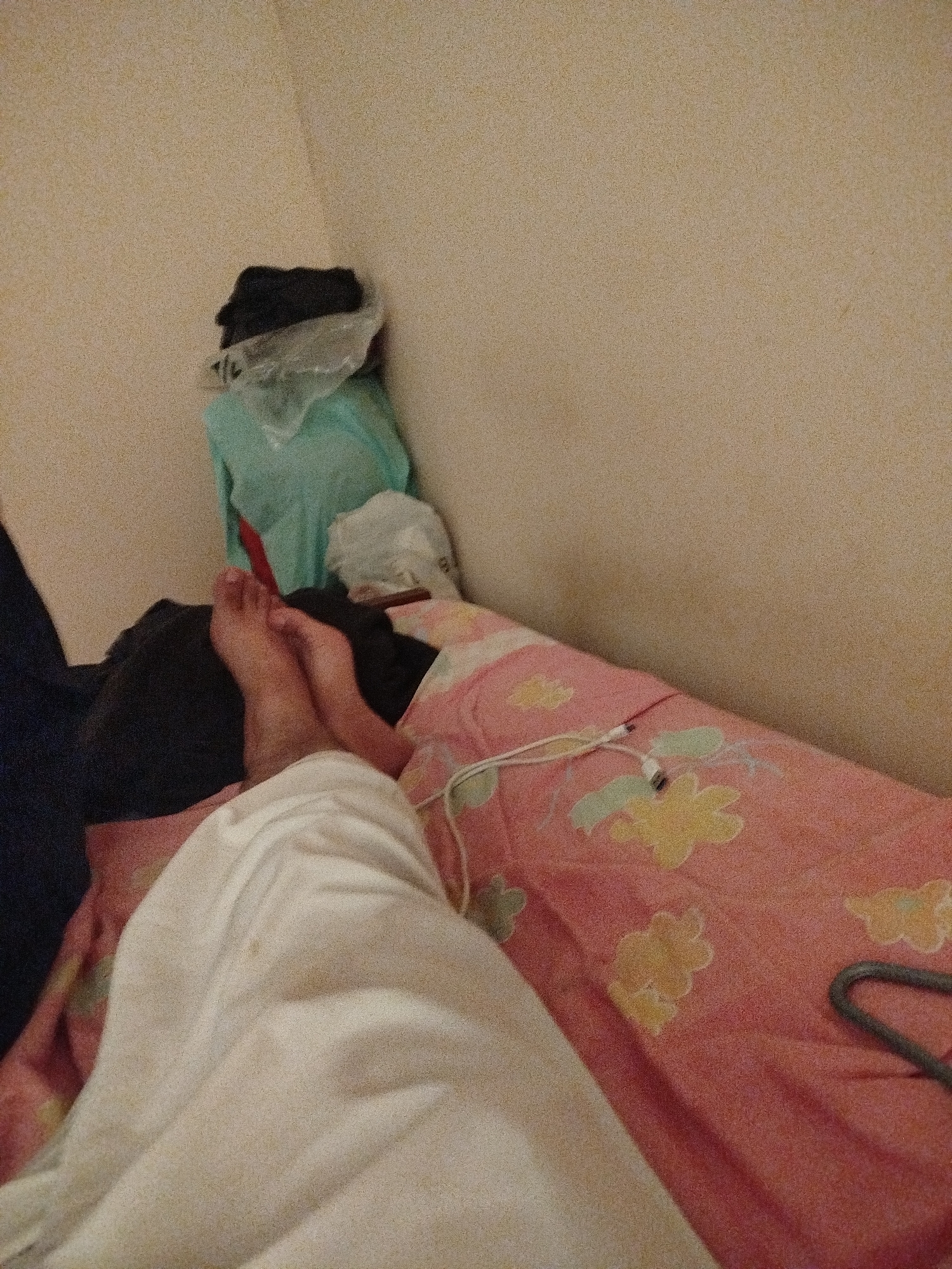 In a dimly lit bedroom, a person's feet are visible resting on a pink and yellow floral comforter. A white sheet is draped over the lower body. Various clothing items are piled on a bed in the background, including a teal garment and dark clothing wrapped in plastic. The room appears cluttered, with a beige wall as the backdrop.