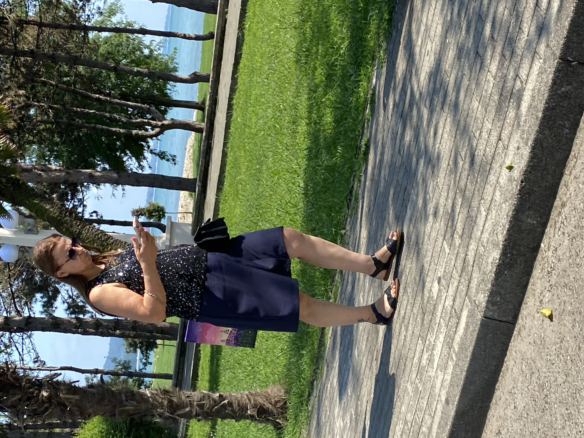 A woman wearing sunglasses and a sequined sleeveless top with dark blue shorts and black sandals is walking on a paved walkway. She is holding a mobile phone in her hands and looking down at it. She is carrying a black purse over her left shoulder. Beside her, a purple and pink picture is leaning against a palm tree. In the background, there is a grassy area with trees, a body of water, and a clear blue sky. The scene appears to be in a park or promenade along the coast.