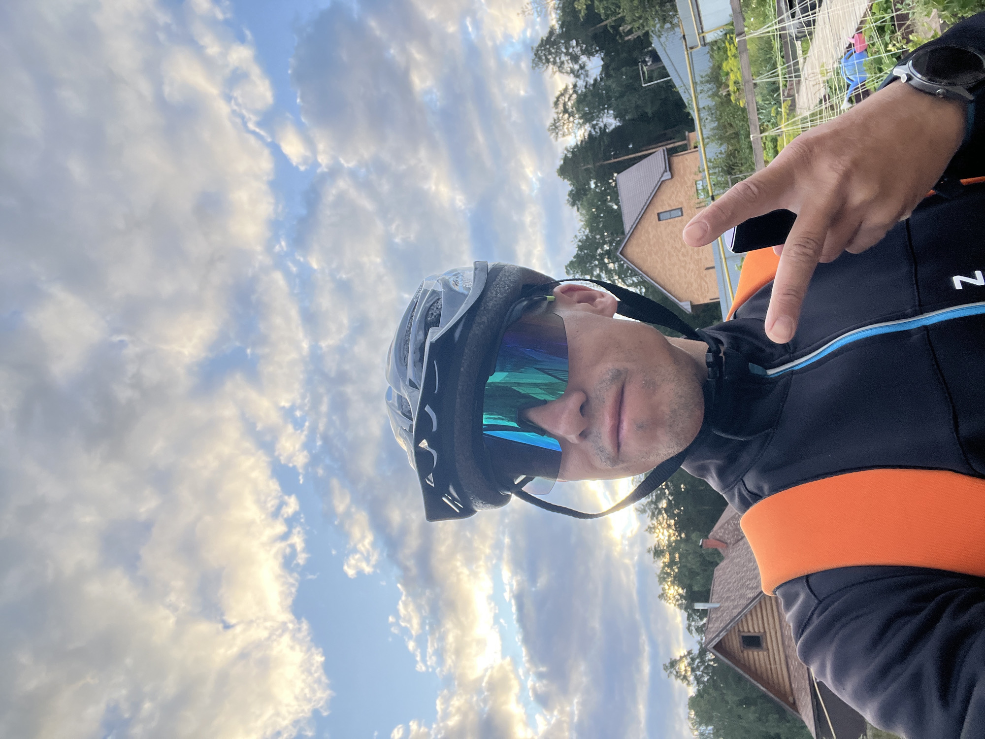 A man wearing a black cycling helmet and mirrored cycling sunglasses takes a selfie outdoors. He is wearing a black jacket with blue and white trim and an orange backpack. His right hand is raised, holding a black smartphone and making a peace sign. In the background, there are trees, houses with wooden and brick facades, and a cloudy sky with patches of sunlight. The image appears to be taken in the city of Chaikovsky, Russia, likely in a suburban or rural area, given the presence of houses and vegetation.