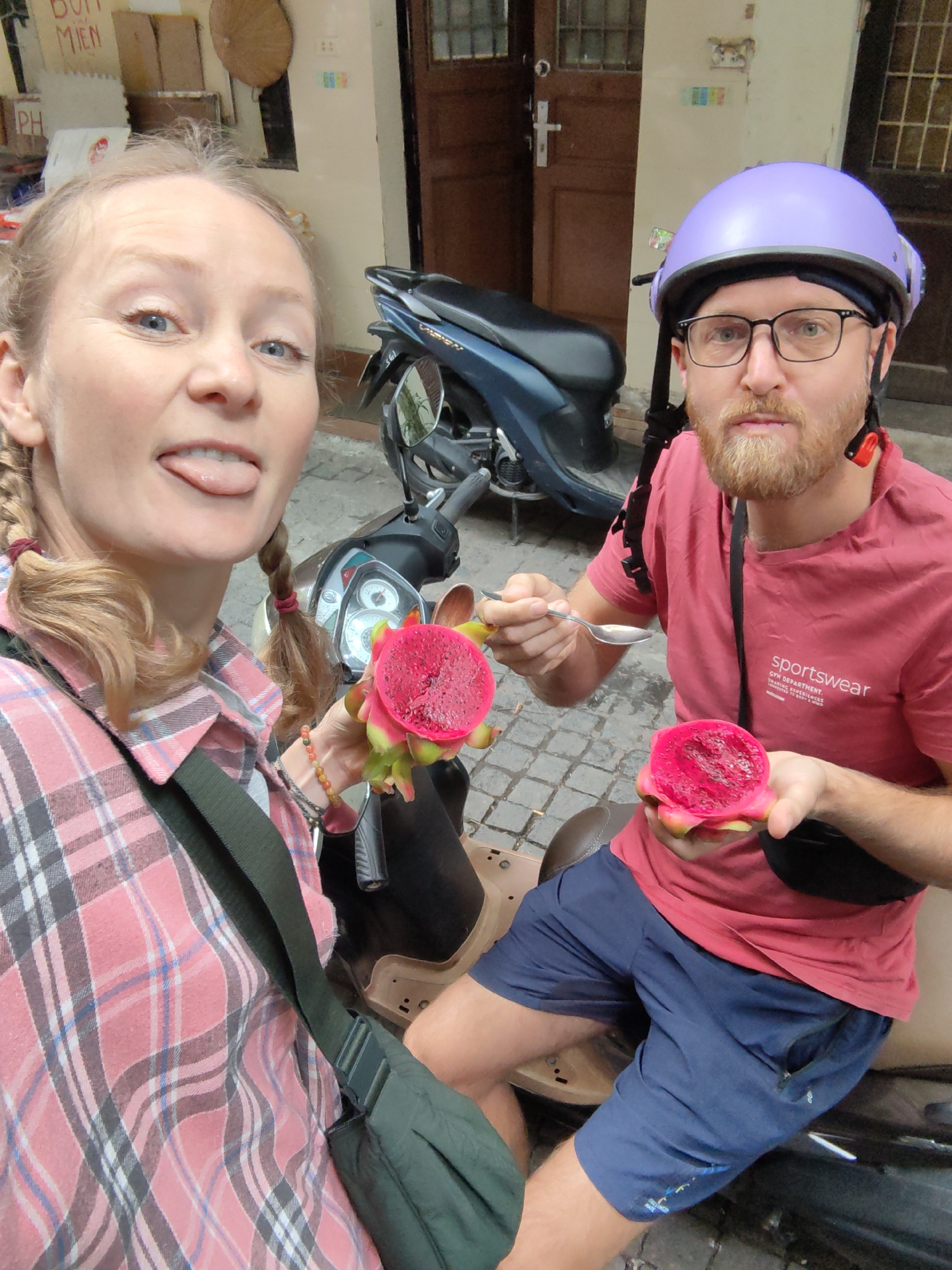 In a vibrant street scene in Hà Nội, Vietnam, a man and a woman pose for a selfie while enjoying red dragon fruit. The woman, with blonde hair styled in two braids, wears a pink plaid shirt and a dark green cross-body bag. She playfully sticks out her tongue towards the camera, holding a vibrant red dragon fruit half.
Beside her, a man with a reddish-blond beard and glasses wears a purple helmet over a black headband, a red t-shirt, and blue shorts. He holds a spoon poised above his dragon fruit half, with another half in his other hand, looking directly at the camera. The red dragon fruit, with its bright pink flesh and small black seeds, is a central focus for both subjects.
Behind them, a blue scooter is parked, indicating an urban setting. The background features light-colored building walls, a wooden door, and a window with a grid pattern. A straw hat hangs on the wall. Visible text on the wall includes "BÚN MIẾN" (a type of Vietnamese noodle dish) and "PH," likely part of "PHỞ." The man's red t-shirt displays the text "sportswear," "GYM DEPARTMENT," "TRAIN YOURSELF," "OVERCOME EVERYTHING," and "MOMENT TO BE GOOD." The ground appears to be paved with cobblestones. The bright lighting suggests it is daytime. The overall mood is casual and cheerful, capturing a moment of enjoyment during their travels.