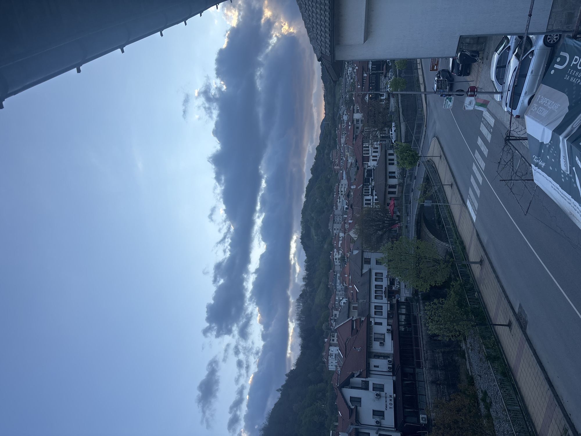 The image depicts an elevated view of a street in Zlatograd, Bulgaria, on a day with dramatic cloud formations.
The sky occupies the upper portion of the frame, showing a mix of blue sky and long, dark storm clouds illuminated along their edges by the setting sun.
Below the sky, the foreground presents a residential area nestled against a verdant hillside. The architecture is dense, with closely packed houses, many featuring red-tiled roofs.
A road runs parallel to the houses. Several parked cars are visible. The road is marked with a pedestrian crosswalk and signs in Bulgarian. A banner ad for "PULZE" is also visible.
Overall, the scene captures a mix of natural beauty and urban landscape in the Bulgarian town.