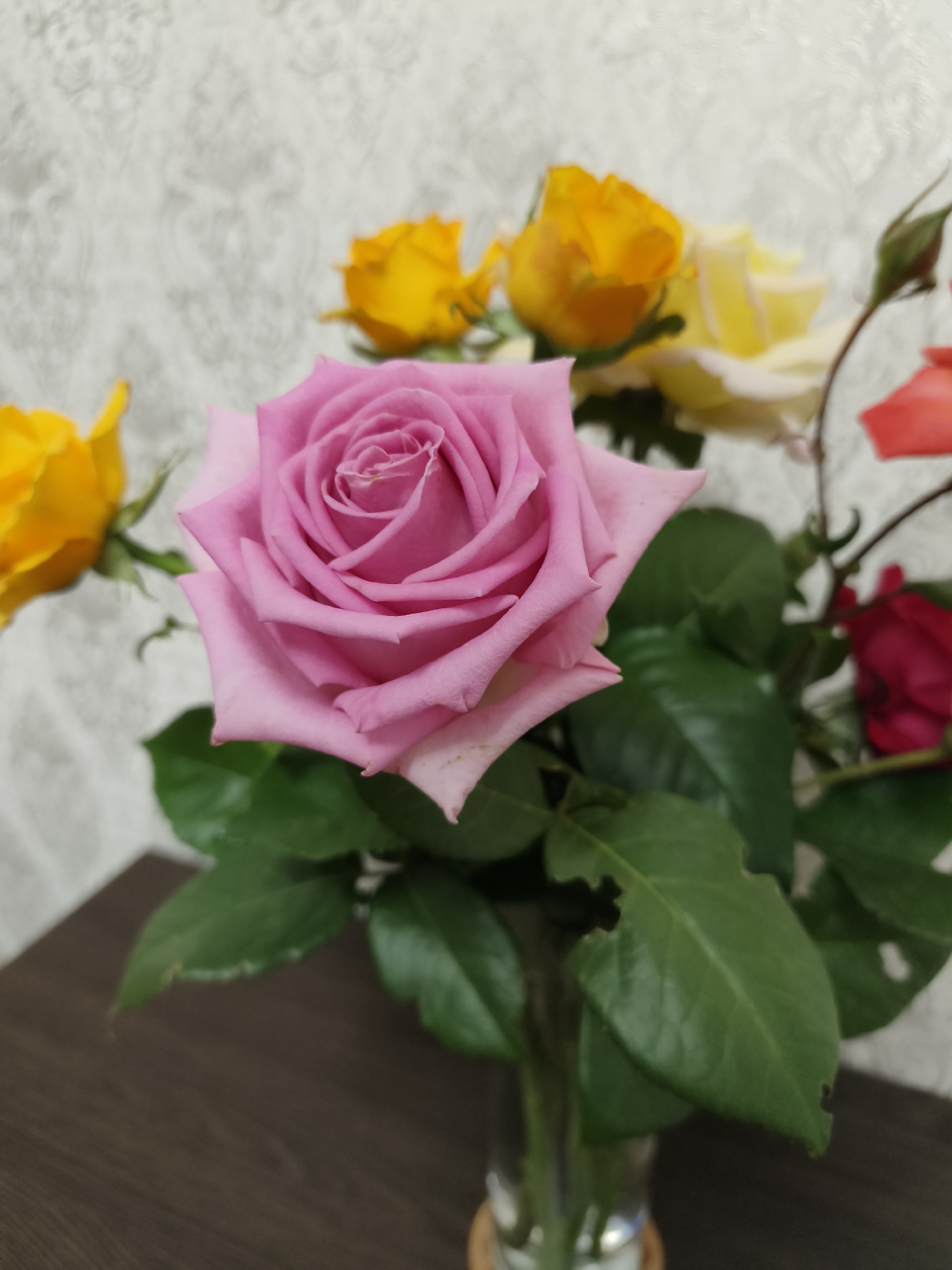 A bouquet of roses is arranged in a glass vase on a dark wooden surface. The most prominent rose is a large, soft pink bloom in the foreground, its petals tightly furled in the center and gently unfurling outwards. Behind it, to the left, is a yellow rose, slightly out of focus. Further back, other roses in shades of yellow, pale yellow, red, and a rosebud are visible. The roses are surrounded by lush green leaves, some of which show small, irregular holes, possibly from insect damage. The background is a wall with a textured, damask-like pattern in a light, neutral color. The lighting is soft, casting subtle shadows and highlighting the delicate textures of the rose petals and leaves. The scene appears to be indoors, on a table or shelf. There is no indication of the location being in Kryvyi Rih, Ukraine, or any other specific city.
