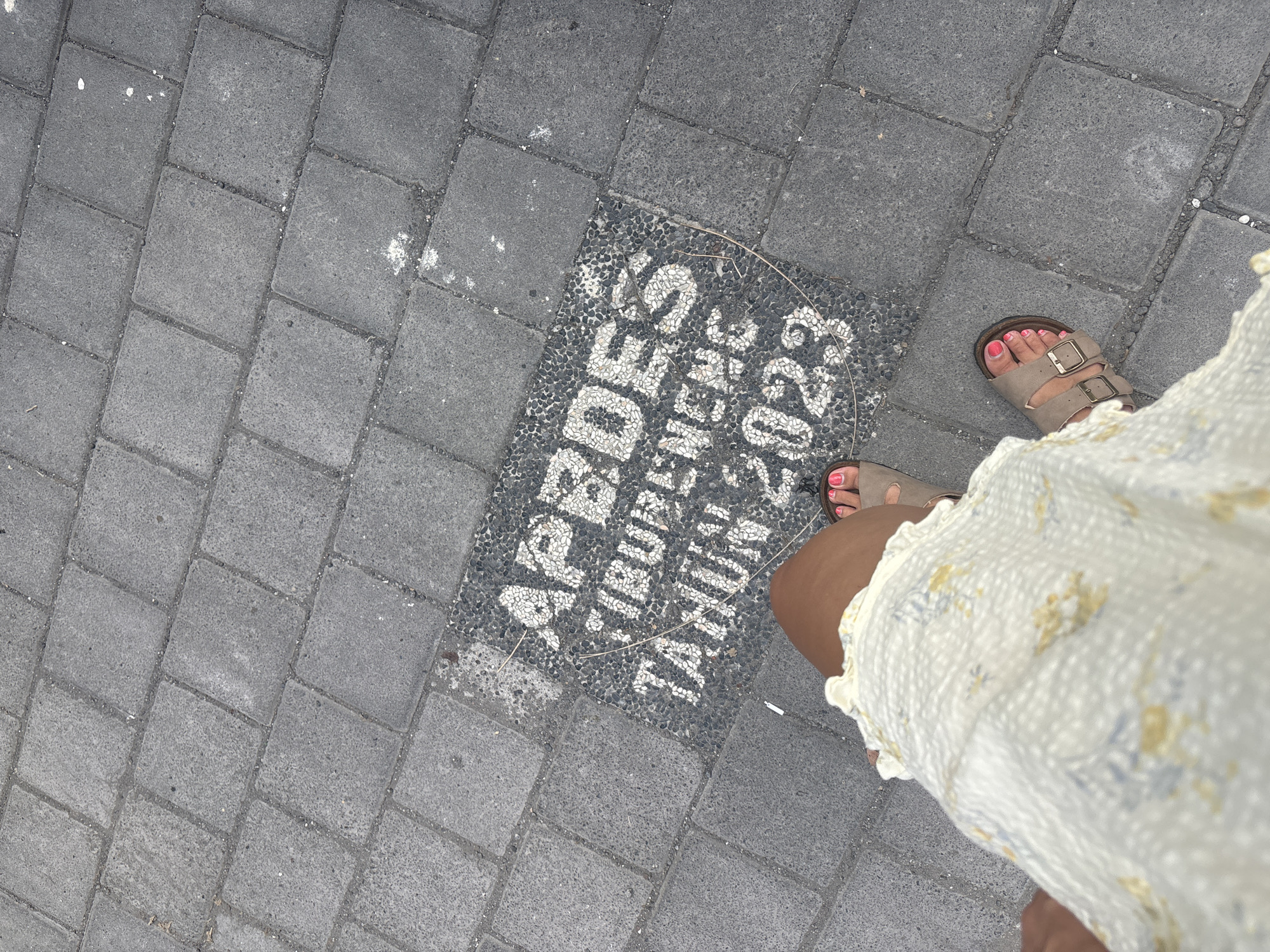 The image shows a section of a paved walkway in North Kuta, Indonesia. The pavement consists of gray rectangular stones. One section of the pavement is inlaid with smaller black and white stones arranged to spell out "APBDes TIBUBENENG TAHUN 2023" in white letters. A person wearing tan sandals and a floral-patterned dress is standing partially in the frame, with their feet and the hem of the dress visible near the inlaid inscription.