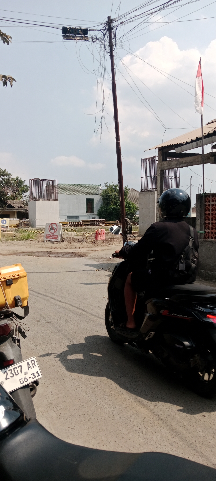 The image depicts a street scene in Purwomartani, Indonesia, with a person riding a black motorcycle in the foreground. The rider is wearing a black shirt, black shorts, sandals, and a black helmet. They are carrying a backpack on their right shoulder. The motorcycle has a yellow cargo box attached to the rear.
In the background, there is evidence of ongoing construction, with rebar structures and scaffolding visible. A white building with a green tiled roof is present, and further construction materials are scattered around. A red and white Indonesian flag is flying from a pole. A sign with a red circle and a diagonal line over a car symbol indicates that vehicles are prohibited. The sky is partly cloudy, and the overall lighting suggests it is daytime. The road surface is asphalt. Another motorcycle is partially visible on the left side of the frame, with a license plate reading "23G7 AR G6.31". Overhead, a complex network of electrical wires is supported by a utility pole.