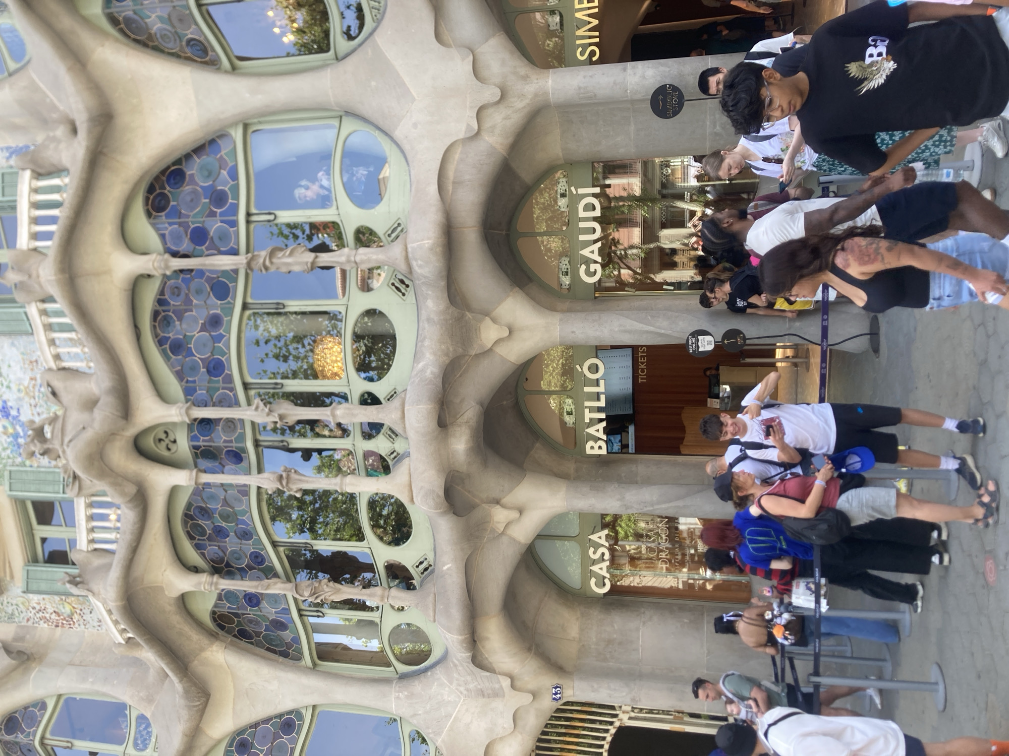The image shows the exterior of Casa Batlló, a building designed by Antoni Gaudí, located on Passeig de Gràcia in Barcelona, Spain. The facade features organic, bone-like structures and colorful stained-glass windows. In front of the building, a queue of tourists is waiting to enter. The people are dressed in casual summer clothing. Some are holding phones, and others are looking around. There are signs indicating "CASA," "BATLLÓ," and "GAUDÍ." The weather appears to be sunny.