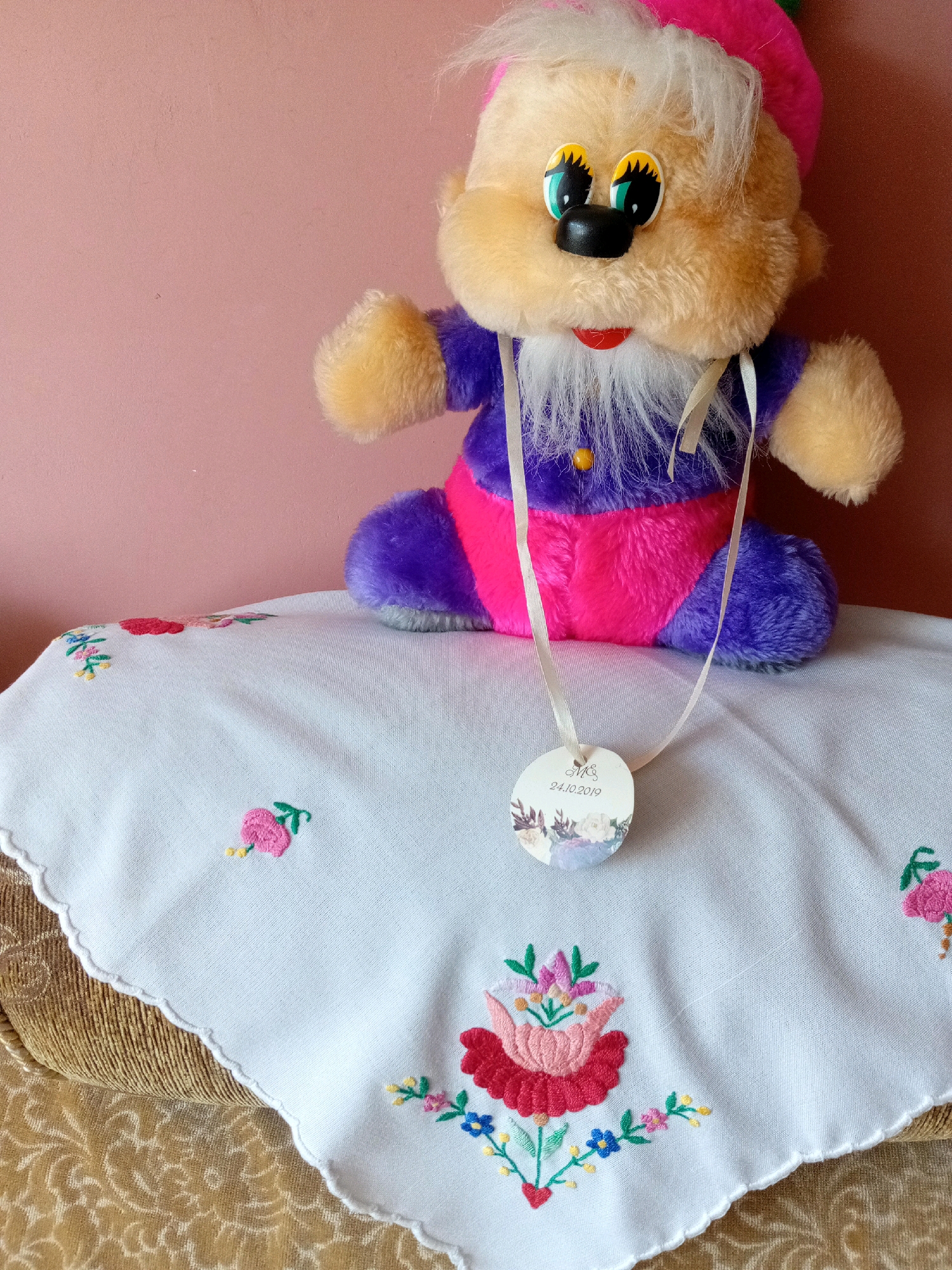 A close-up, eye-level shot captures a plush toy sitting on a white embroidered cloth. The toy, with a beige fuzzy face, big cartoon eyes with green irises and black pupils, a black button nose, and a wide red smile, wears a purple and pink outfit and a pink hat. A white ribbon is tied around its neck, from which hangs a circular tag displaying floral imagery and the text "M&S 24.10.2019". The embroidered cloth, with a scalloped edge, features various floral motifs in pink, red, blue, and yellow. The background is a plain, light pink wall. The scene appears to be indoors, possibly in a home environment in Tiachiv, Ukraine, given the context.