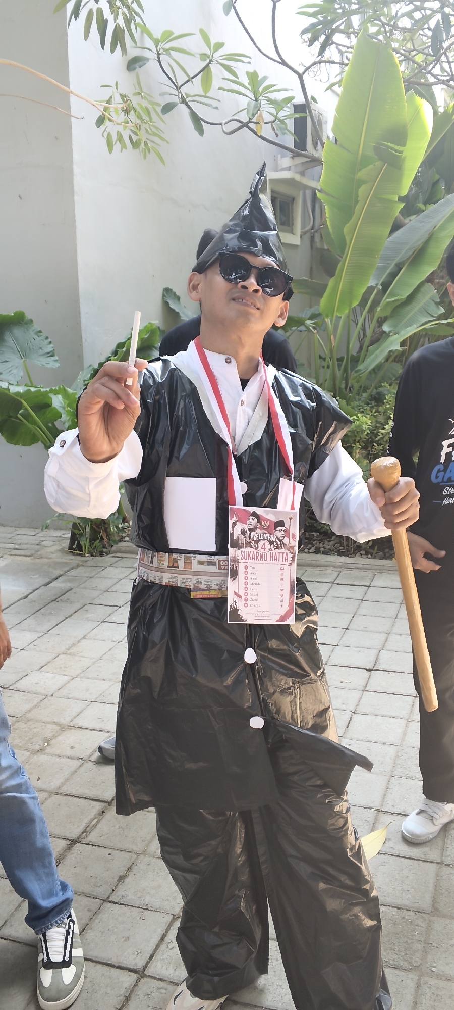 A person dressed in a black trash bag costume, complete with a conical black hat, sunglasses, and a red and white ribbon necklace, stands outdoors in Talise, Indonesia. The individual holds a cigarette in their right hand and a wooden staff in their left. A poster attached to their costume features portraits of two men and the text "KELOMPOK 4 SUKARNO HATTA," along with a list of names and symbols. The person is standing on a paved walkway, with lush green foliage visible in the background, suggesting a park or outdoor event setting.