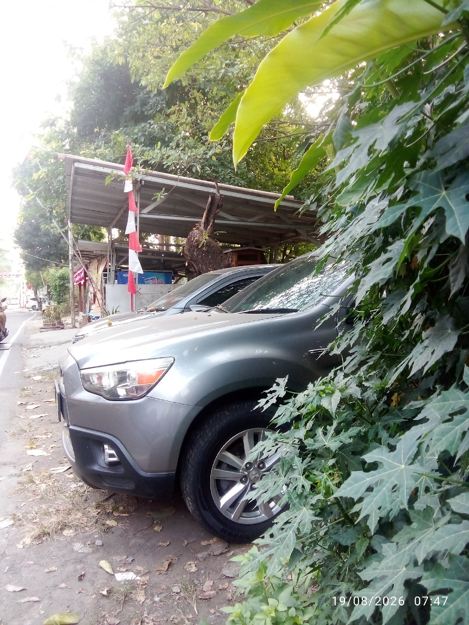 In East Jakarta, Indonesia, a silver SUV is parked on the side of a street. The vehicle is partially obscured by lush green foliage in the foreground. Behind the SUV, a simple open-sided structure with a corrugated metal roof is visible. Red and white Indonesian flags are hung from a pole near the structure. The ground is a mix of dirt and dry leaves. A road with a white line is to the left of the parked car, and trees line the background. The time stamp on the image reads "19/08/2026 07:47".