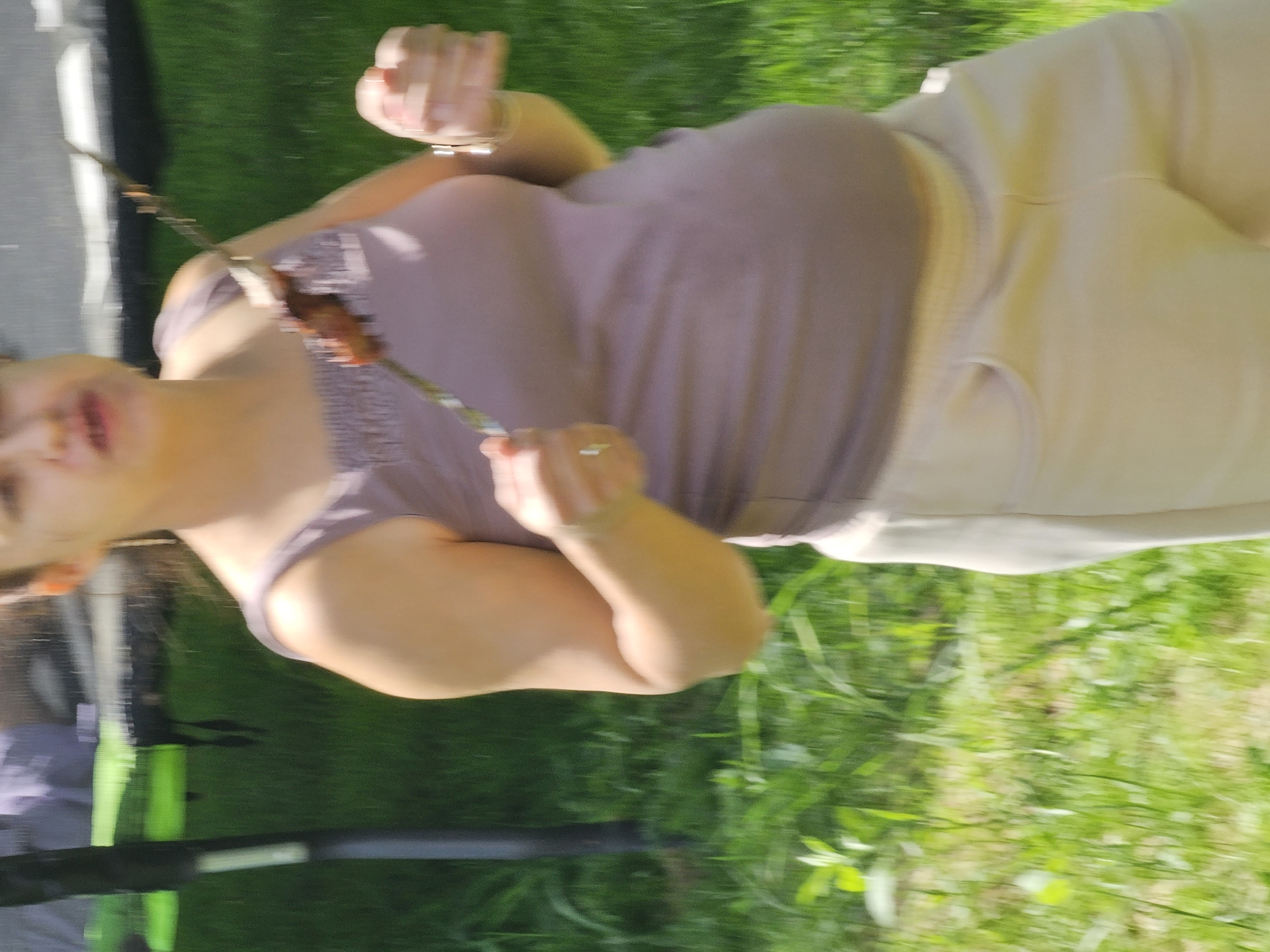 A woman stands outside on grass, possibly in Мышкино, Russia, holding a skewer with food on it. She wears a mauve tank top and light-colored pants. The background is green grass. Part of an object that appears to be a baby stroller is visible to the left of the frame.