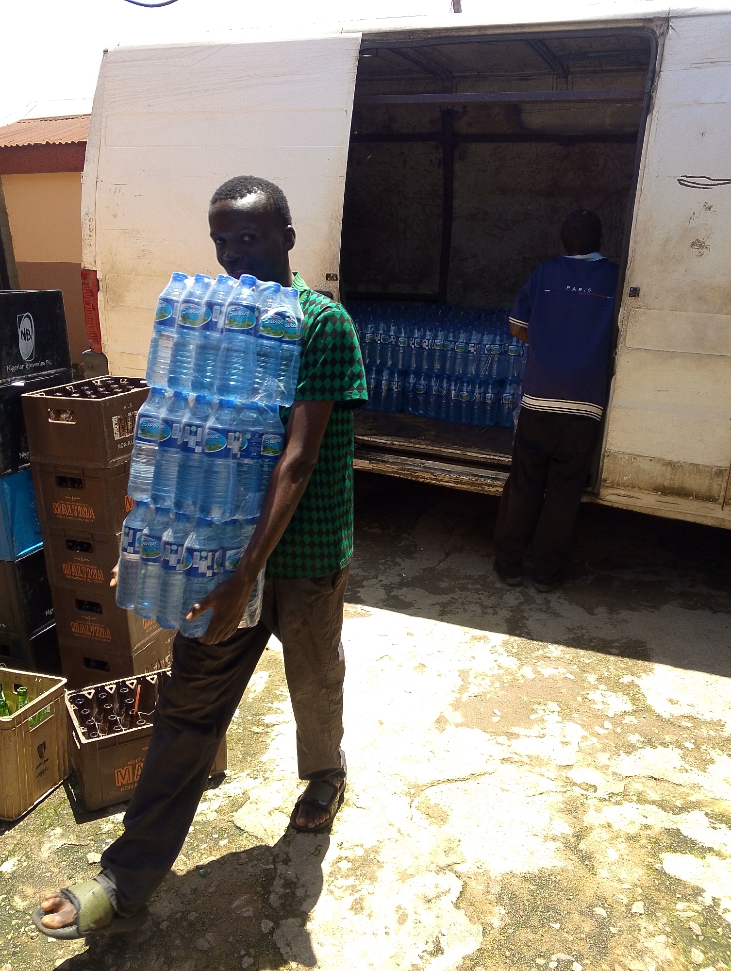 A Black man is standing in front of a white truck, carrying a large stack of bottled water. He is wearing a green and black checkered shirt and brown pants. The truck's back door is open, revealing rows of bottled water inside. There are also stacks of crates with drinks outside the truck. The ground is paved and shows signs of wear and tear. The scene appears to be in an outdoor urban setting.