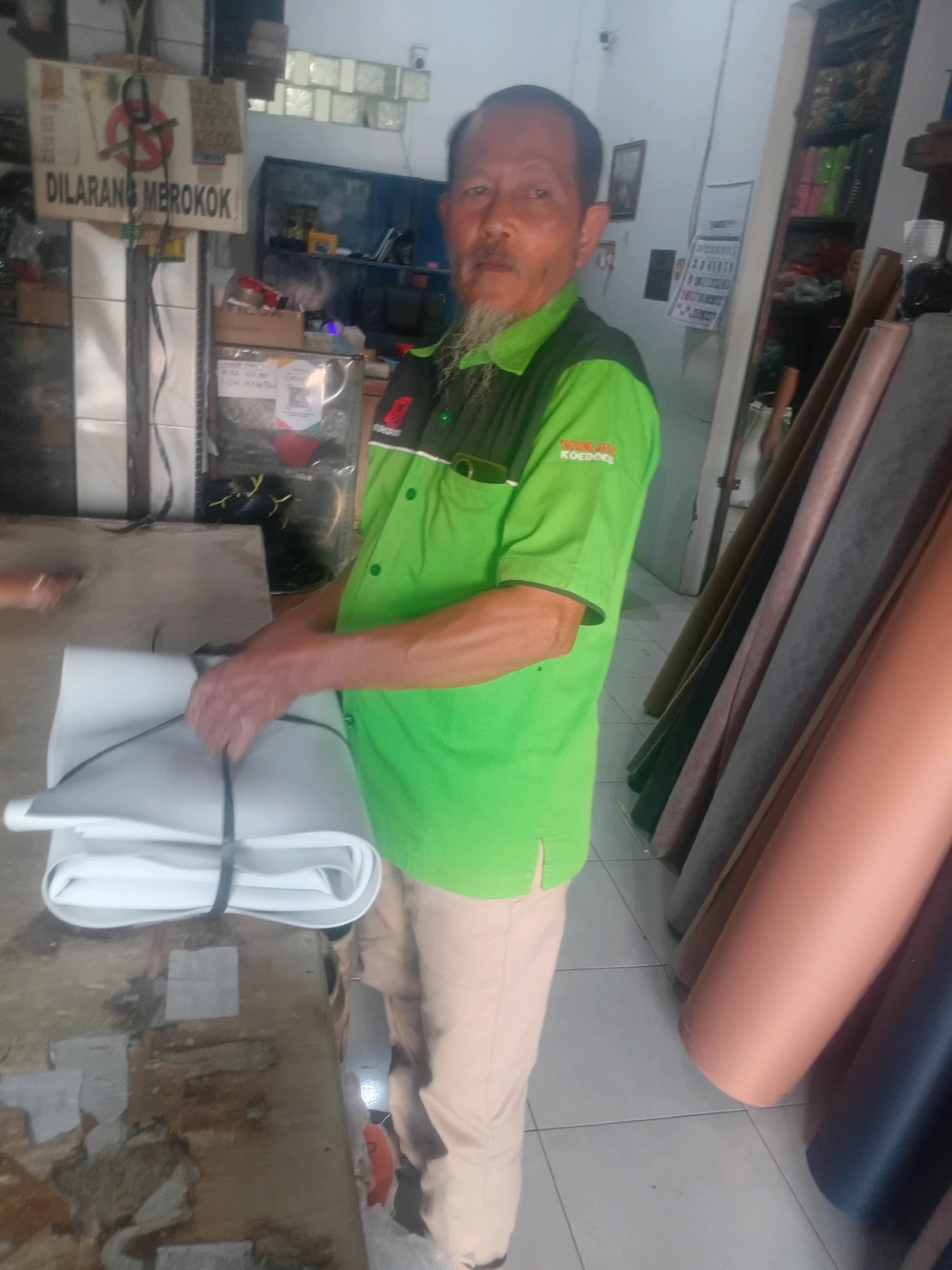 In Kerjasan, Indonesia, a man in a bright green polo shirt with a "KOEDOES" logo stands in a workshop. He has a mustache and a goatee and is looking at the camera. He is holding a large, rolled-up white material, possibly fabric or leather, tied with a black strap. In the background, there are shelves filled with various items, including what appear to be rolls of material in different colors and textures, leaning against a wall. A "DILARANG MEROKOK" (No Smoking) sign is prominently displayed on a pole to the left of the man. The floor is tiled, and the overall environment suggests a commercial or workshop setting.