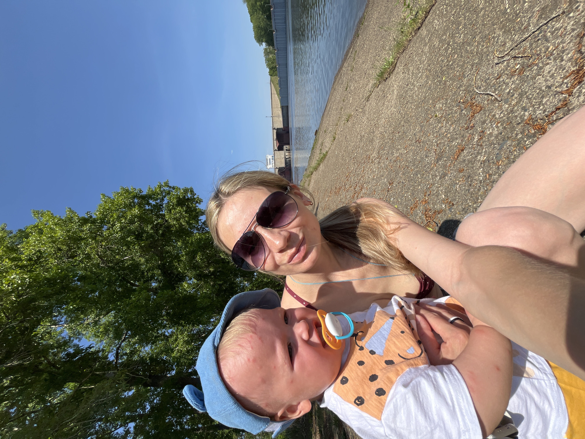 The image is an outdoor selfie taken in Chaikovsky, Russia, featuring a woman holding a baby. The location appears to be near the Kama River, with a concrete embankment visible on the right, and a dam or industrial structure across the water.
The woman is wearing sunglasses and a swimsuit top, suggesting warm weather. She has blonde hair. The baby is wearing a blue hat and a shirt with a fox-like cartoon design and has a pacifier in their mouth.
The background includes a clear blue sky, a large green tree, and the Kama River with industrial structures in the distance. The lighting indicates it's daytime. There are no explicit actions occurring, but the woman is holding the baby, and they are both facing the camera.