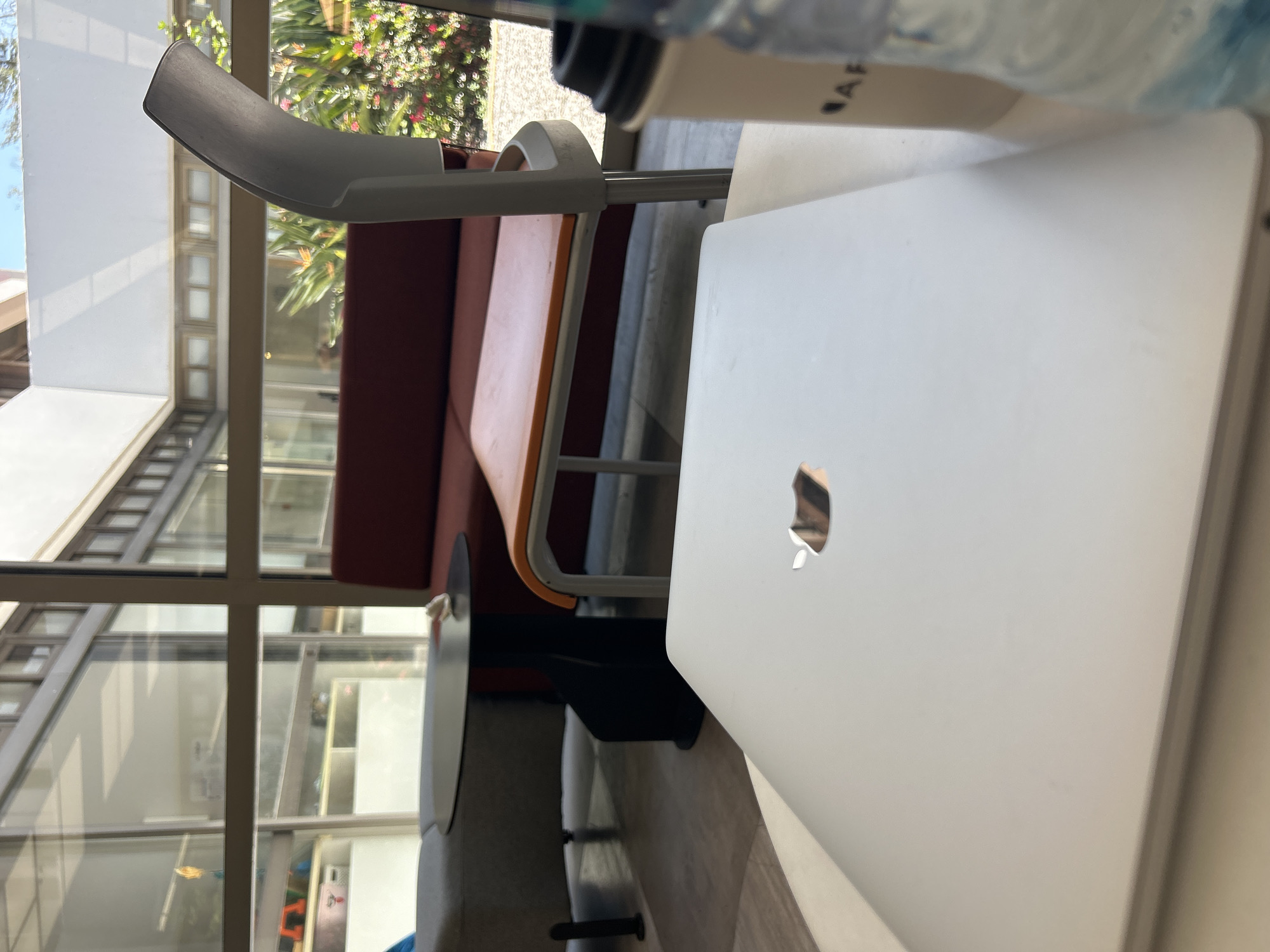 The image displays an indoor setting, possibly a cafe, lounge, or study area, with a large window offering a view of the outdoors. The image is oriented vertically, but depicts a horizontal scene.
In the immediate foreground, a silver MacBook laptop is prominently featured on a light-colored surface, positioned to the right side of the frame. Its distinctive Apple logo is clearly visible on its closed lid. To the upper right of the laptop, parts of a white paper coffee cup and a clear plastic water bottle are visible. The letters "AF" can be seen printed on the side of the coffee cup.
The midground includes modern furniture. A light grey and white chair with a curved backrest is positioned towards the center-left. Behind it, a long, reddish-brown upholstered bench or couch stretches horizontally across the frame. A dark, round table is partially visible to the right of the chair and in front of the couch.
The background is dominated by expansive windows that look out onto a bright, sunny day. Outside, vibrant green foliage is visible, interspersed with clusters of small pink or red flowers, suggesting a lush, warm environment. Reflections in the window also show parts of an interior building structure or hallway with multiple window panes, indicating another section of the building or a nearby structure.
The scene appears tranquil, filled with natural daylight. No people are present. The time of day is clearly daytime, given the brightness.
