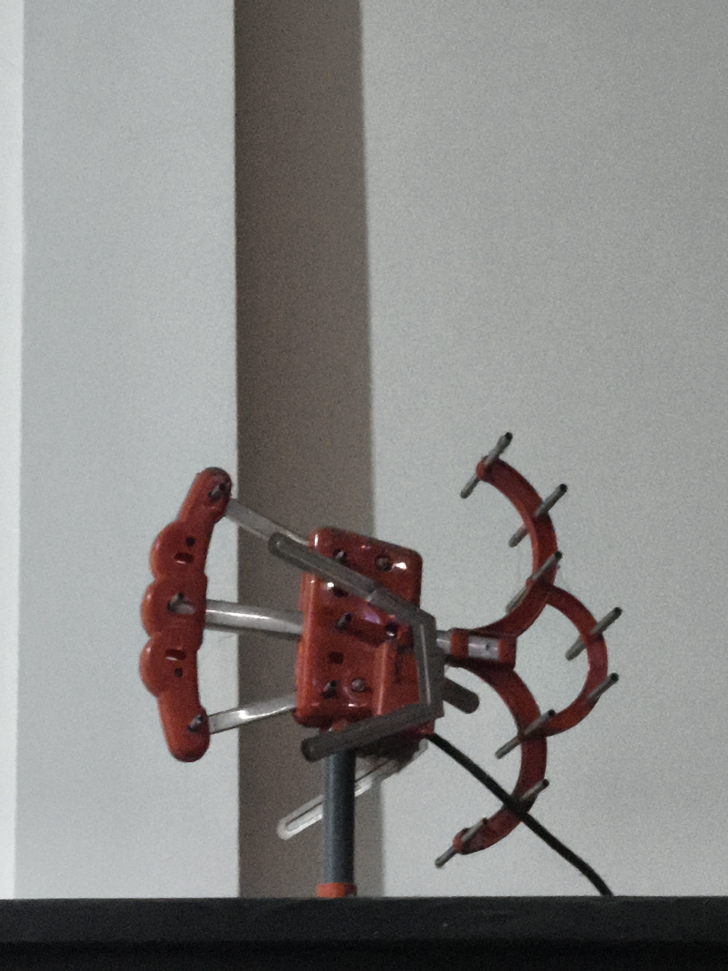 The image shows a close-up of a red and silver TV antenna with several prongs. The antenna is mounted on a grey pole with an orange cap at the bottom, which rests on a dark surface. The background is a plain, light-colored wall with a vertical shadow running down it. There are no people or animals visible in the image. The scene is indoors, likely in a residential or commercial setting.