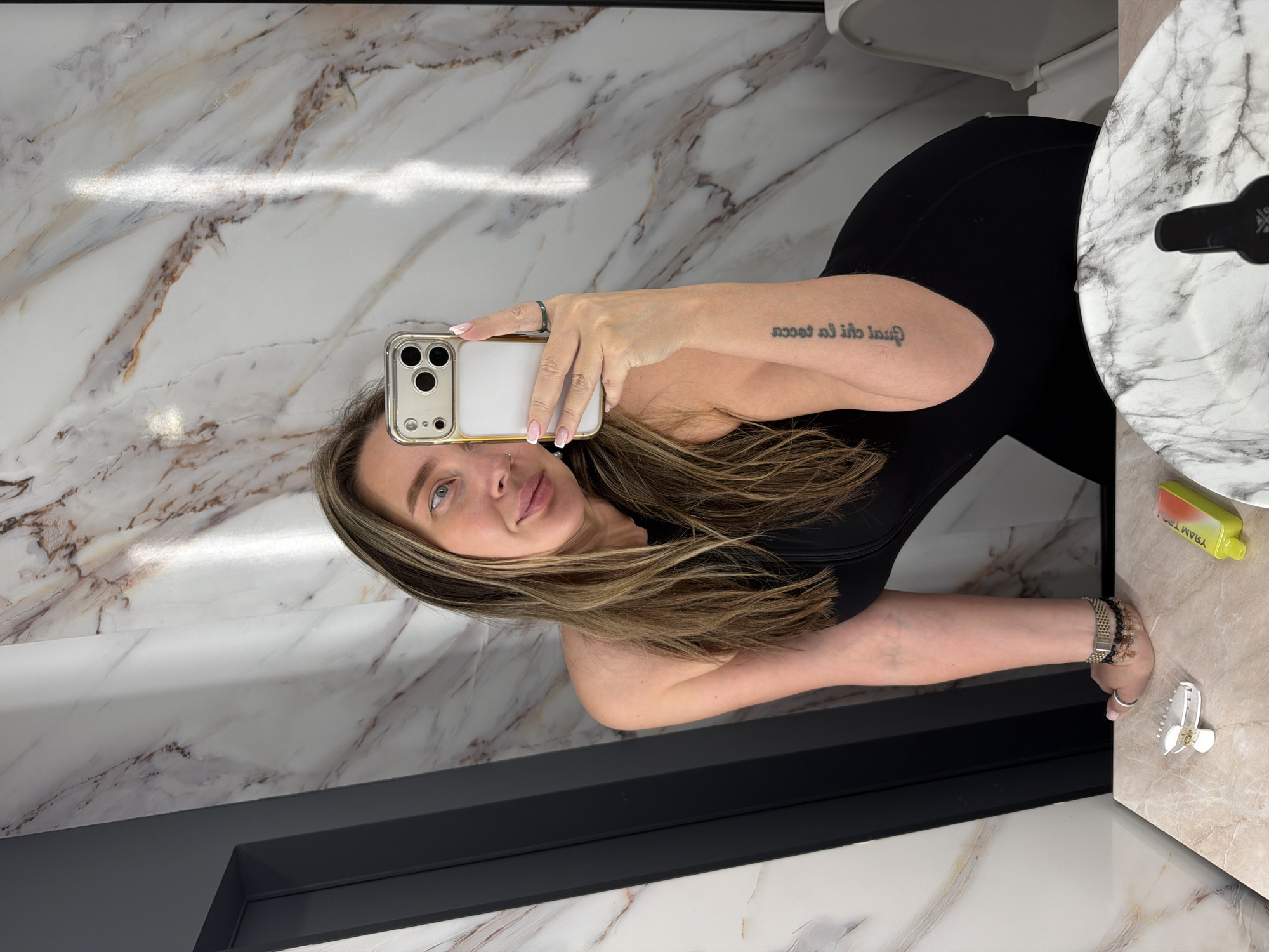 A woman is taking a selfie in a bathroom with marble walls and flooring. She is wearing a black top. Her hair is blonde with dark roots. She is holding a white and gold iPhone. There is a sink and a toilet visible in the background. A bottle and a hair clip are visible on the sink counter. The location cannot be identified as Moscow, Russia, from the provided image.