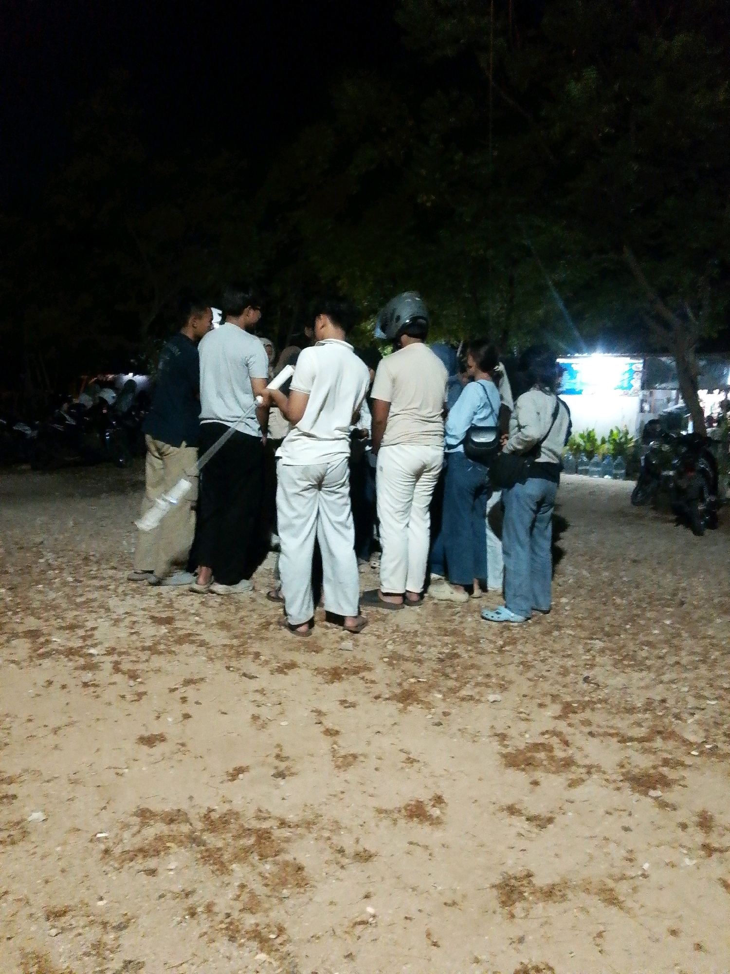 A group of ten individuals, appearing to be young adults, are gathered outdoors at night in what seems to be an open, dirt lot in Cikal, Indonesia. The group is clustered together, with most facing inward toward the center of the group.
Several individuals are wearing casual attire. One person in the foreground on the right is wearing light-colored pants and a light-colored shirt, standing with their back to the camera. To their left, another person is wearing dark pants and a dark top, and is facing away from the camera. Further left, a person in a light-colored shirt and dark, possibly sarong-like, pants is partially obscured. A person in a light-colored shirt and light-colored pants is visible in the center of the group. Another individual in a light-colored shirt and dark pants is seen on the far left.
Among the group, one person is wearing a dark helmet. Two women are also present, one in a light blue top and dark pants, carrying a bag on their shoulder, and another in a light-colored top and dark pants, also with a bag.
The environment suggests a rural or semi-rural setting. The ground is covered in dirt and scattered debris. In the background, trees with dark foliage are visible, indicating a wooded area. Further back, there are lights from what appears to be a building or stalls, and several motorcycles are parked. The overall lighting suggests artificial light sources illuminating the scene. The atmosphere appears to be relaxed and social.