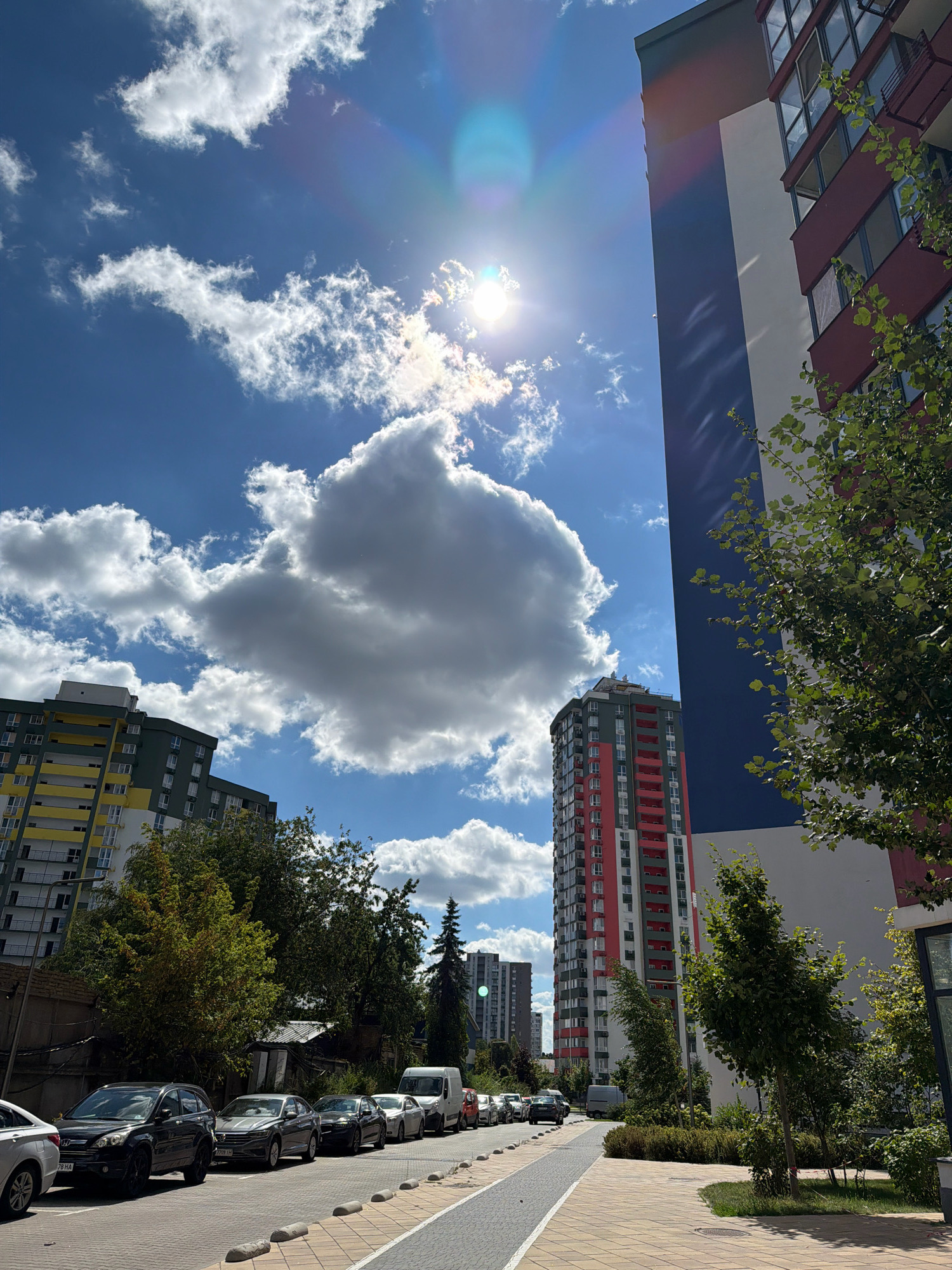 The image captures a sunny day in Kyiv, Ukraine, featuring a street lined with parked cars and modern apartment buildings. The sky is a vibrant blue, dotted with fluffy white and grey clouds, with the sun prominently visible and casting lens flare. A tall apartment building with a distinctive red, white, and blue facade stands on the right, partially obscuring a similar but taller red and green building further down the street. To the left, another apartment building with yellow and green accents is visible. The street is paved with cobblestones and asphalt, with a pedestrian walkway bordered by manicured landscaping. Several cars, including a black SUV, are parked along the roadside. The overall scene depicts a contemporary urban environment with clear weather.