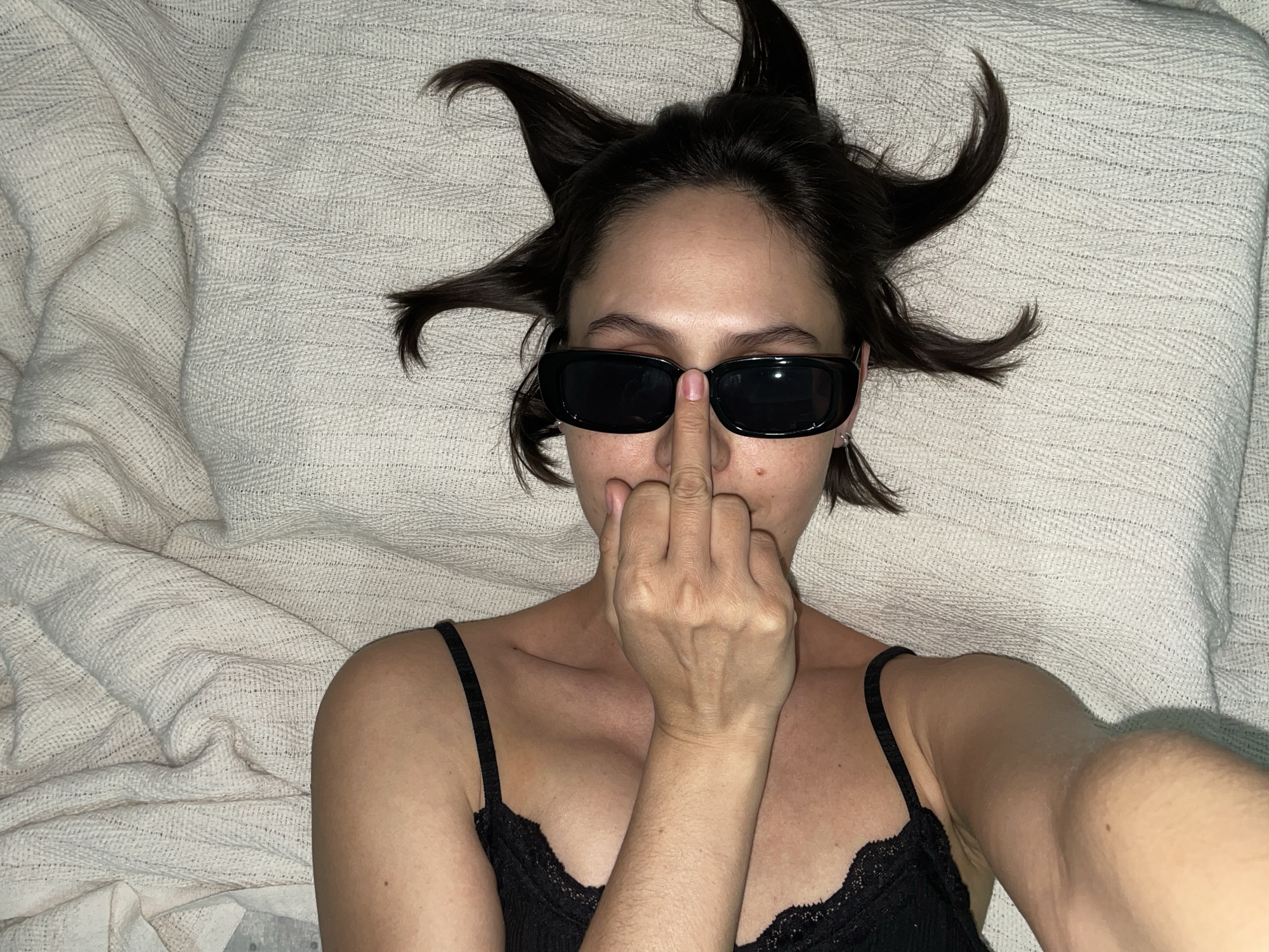 A person wearing dark sunglasses and a black lace-trimmed camisole lies on a textured, light-colored surface. The person's dark hair is styled in a way that projects upwards and outwards. The person is extending their middle finger towards the viewer, with the finger positioned near the sunglasses. The scene appears to be indoors. There is no indication of Izhevsk, Russia, in the image.