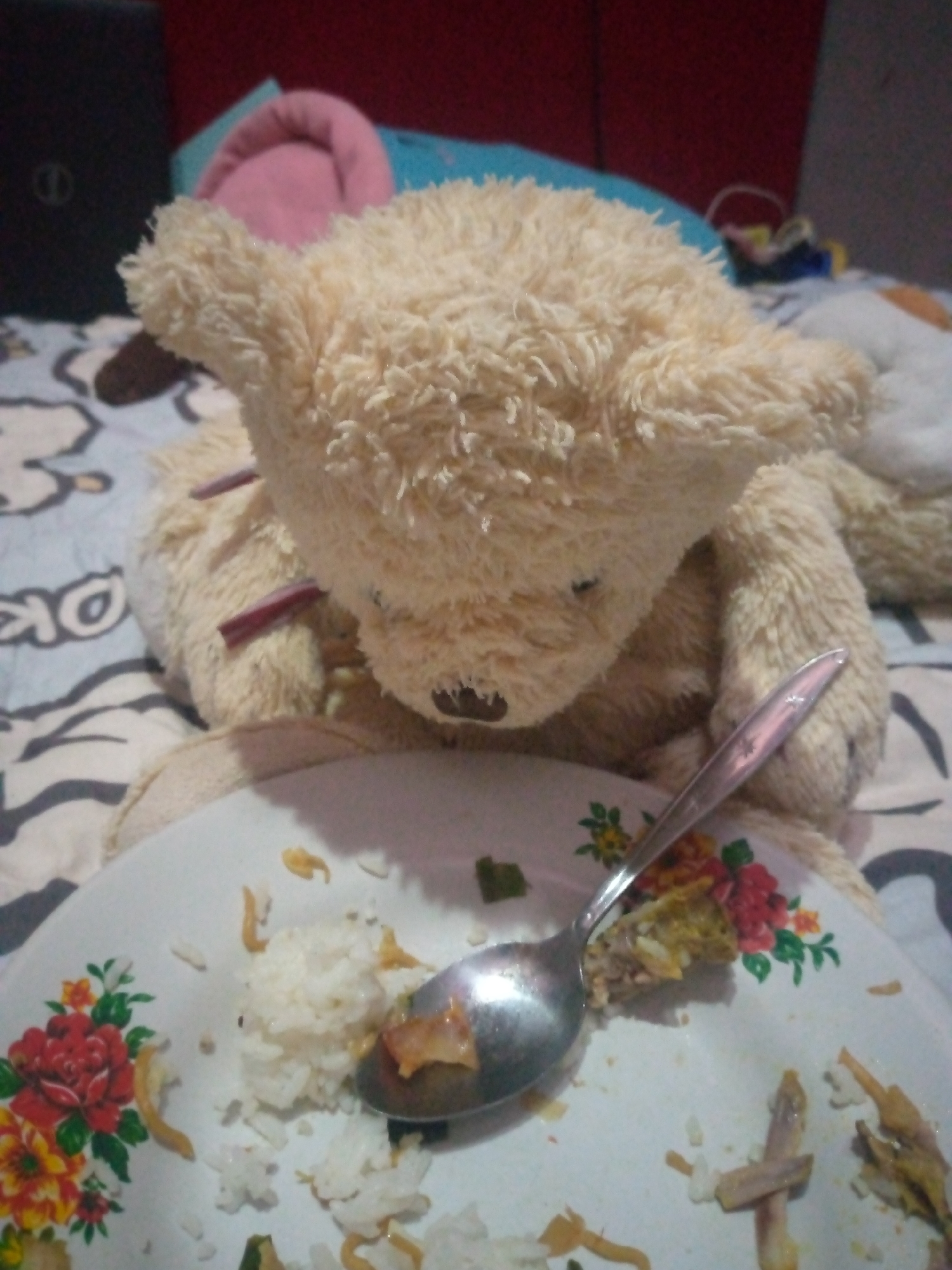 A light brown teddy bear is positioned on a bed, leaning against a red headboard. A plate of rice and chicken is in front of the bear, with a spoon in the plate. The scene is indoors, likely in a bedroom.