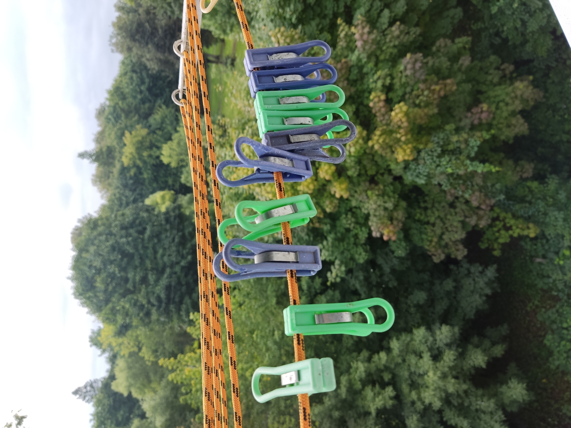 A row of green and blue clothespins are clipped onto two parallel orange and black ropes, suspended horizontally against a blurred backdrop of lush green trees and a cloudy sky. The ropes are attached to white metal fixtures. Two green clothespins are on the left, followed by a cluster of green and blue clothespins in the center and right. The scene appears to be outdoors, likely on a balcony or clothesline, with a view overlooking a densely wooded area, characteristic of a park or forest on the outskirts of a city. The image has a depth of field that blurs the background trees, drawing focus to the clothespins. The lighting suggests an overcast day.