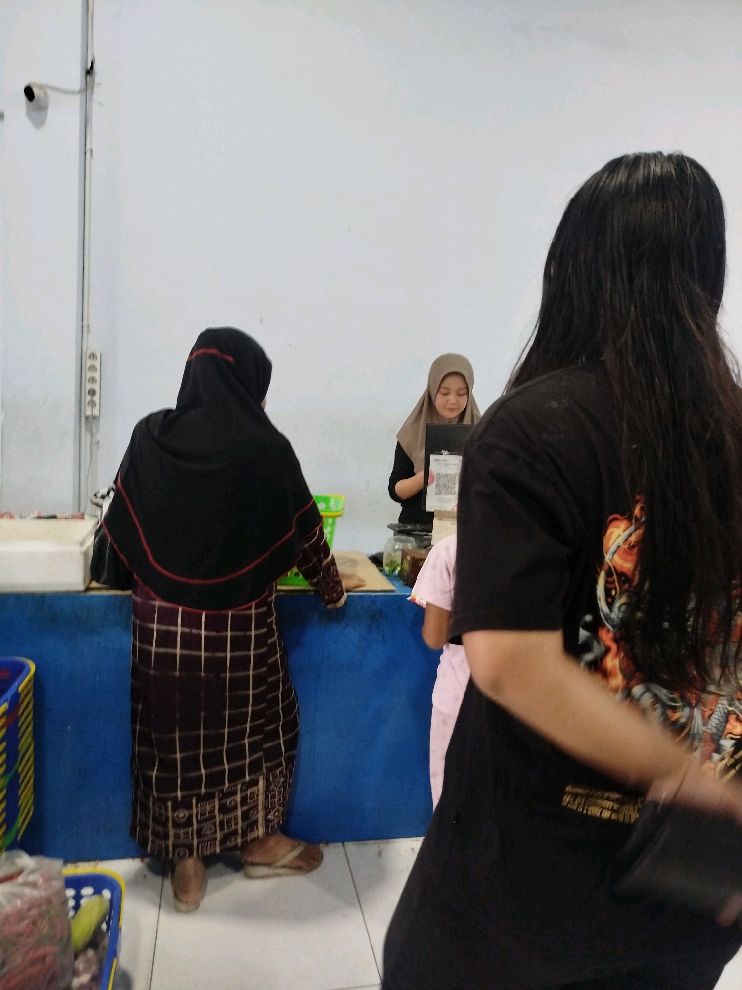 A medium shot captures a scene likely taking place in a market or shop in Kadipaten, Indonesia. In the foreground, a person with long, dark hair is seen from behind, wearing a black t-shirt with a graphic design on the back. They are holding a dark object in their right hand. To the left, a person in a black hijab and a patterned dress is facing away from the camera, standing at a counter. They appear to be interacting with someone behind the counter. Further back, a young person wearing a hijab is behind the counter, looking down at a device that displays a QR code. The environment includes a blue counter, white walls, a security camera mounted on the wall, and some plastic baskets containing goods. The lighting suggests an indoor setting, possibly during the day.