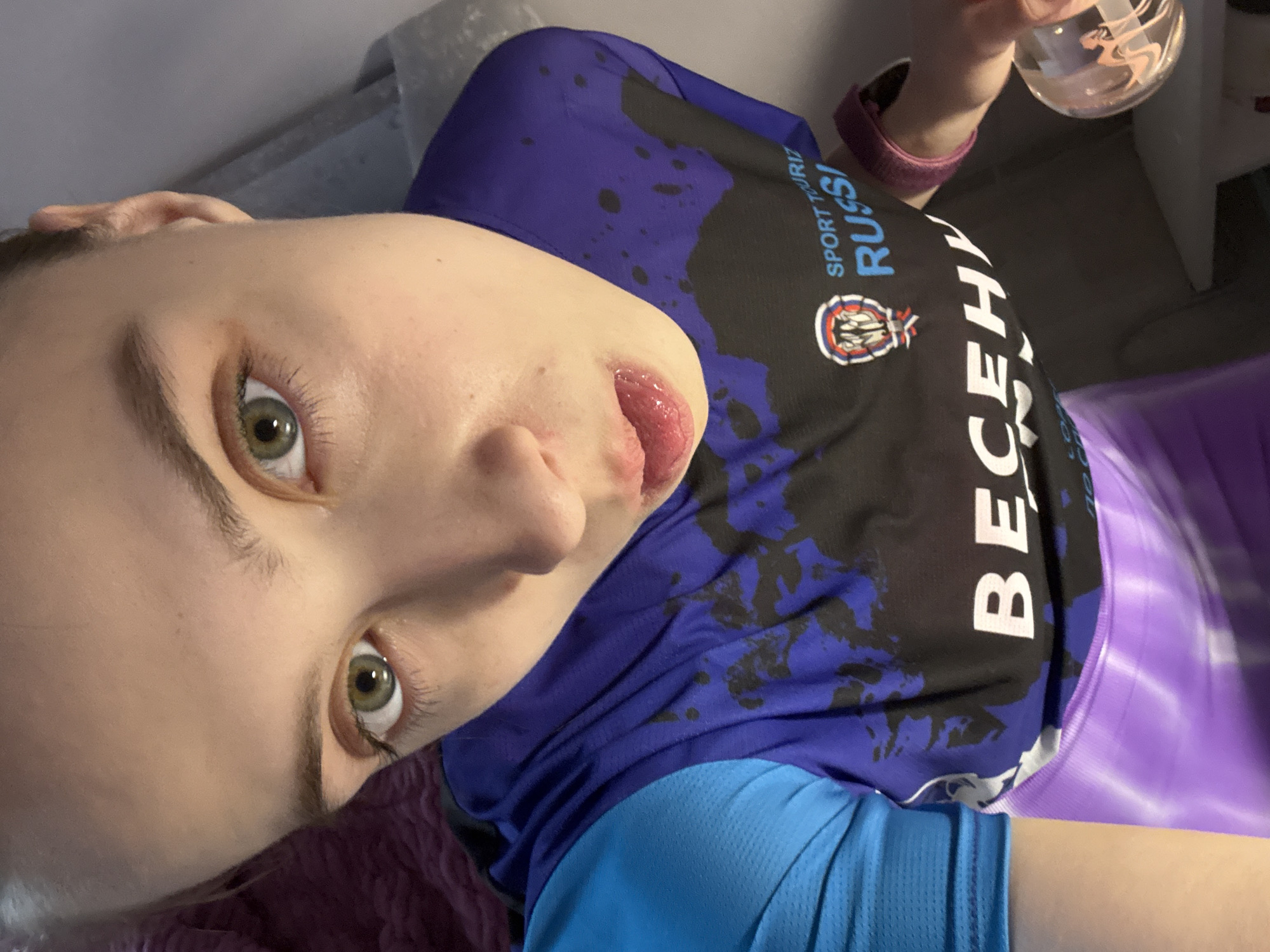 A close-up selfie shows a person with light skin, green eyes, and light eyebrows. The person is wearing a black and blue shirt with the word "RUSSIA" printed on it in white Cyrillic letters. The shirt also has a logo on it. The person is holding a clear glass with a silhouette of a woman. The location of the picture is in Chaikovsky, Russia.