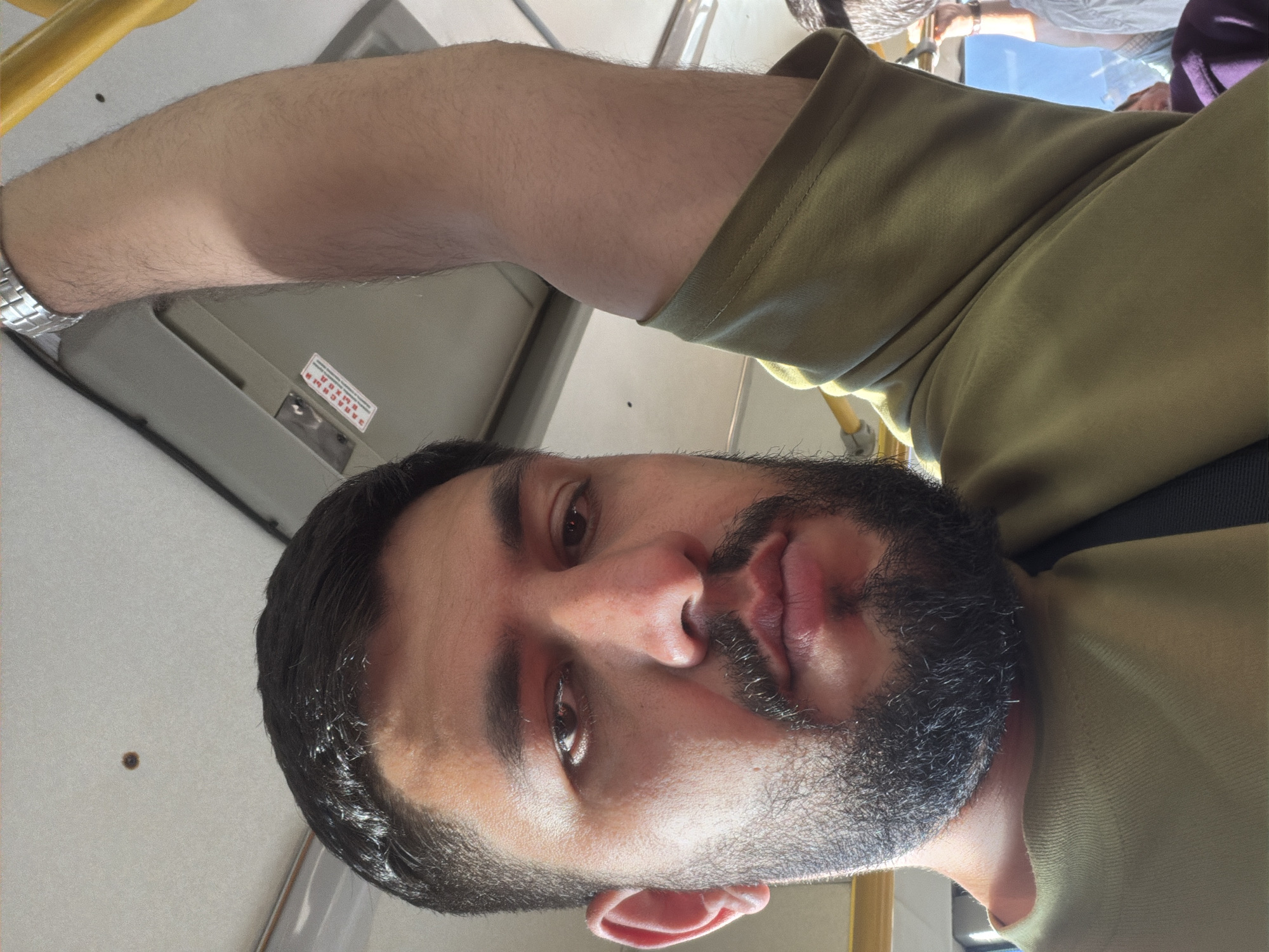 A bearded man takes a selfie. He is on public transportation, possibly a bus, as indicated by the overhead handrail and interior fixtures. He is wearing a light green or olive-colored shirt and a watch on his left wrist. His surroundings include the interior of the vehicle, with a handle, a grey panel above his head and other passengers are visible in the background. There is no readily available information to confirm it is the city of Krasnoyarsk, Russia.