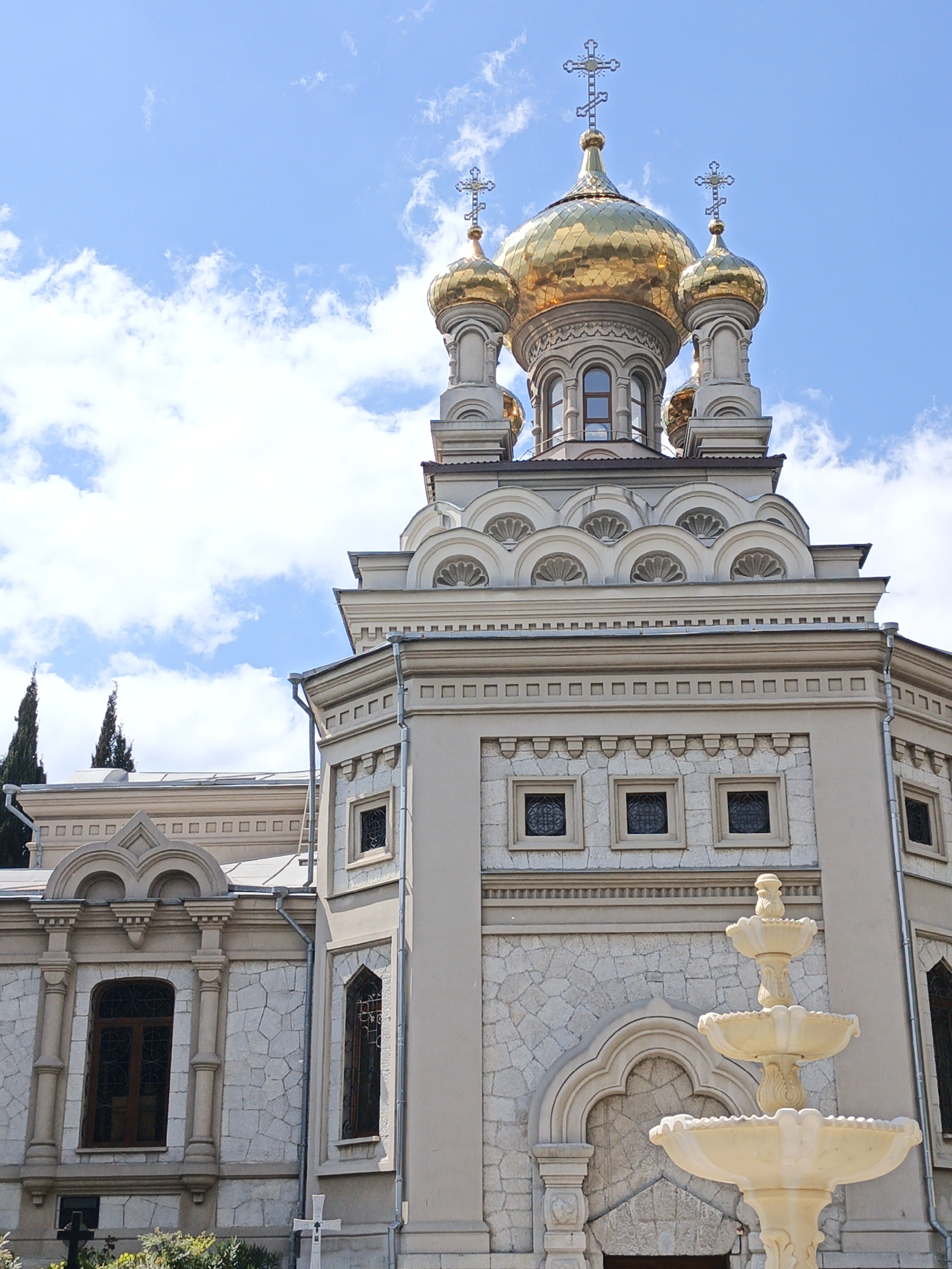 The image shows a tall church structure with golden domes topped with crosses against a partly cloudy blue sky. The church features stone walls with decorative arched windows, and a multi-tiered fountain is in the foreground. The location is Unknown, Unknown.