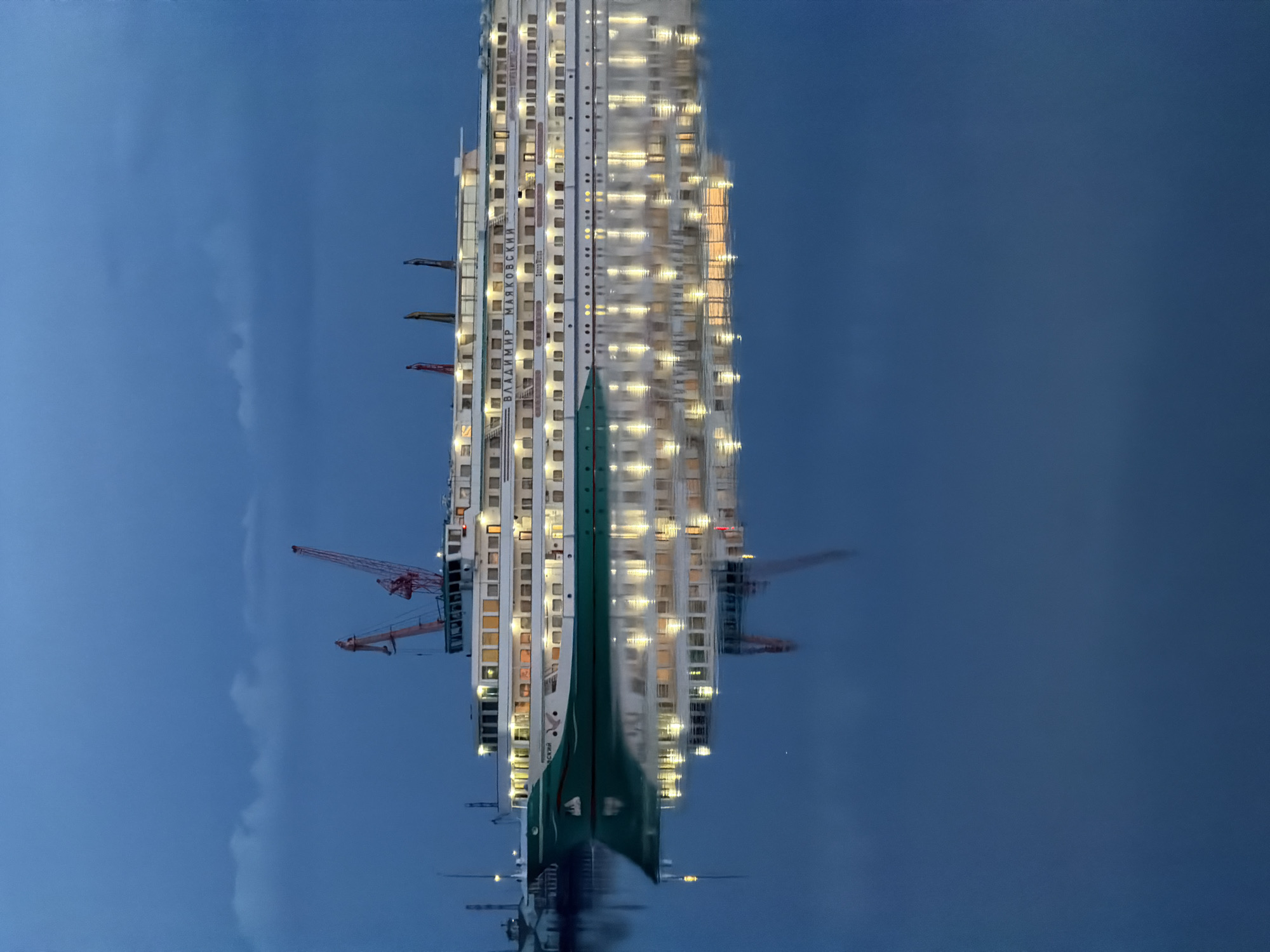 The image shows an inverted view of the "Vladimir Mayakovsky" motor ship reflected in the water. The ship has a tall, multi-deck structure with numerous illuminated windows. The name of the ship is visible in Cyrillic script on its side. A construction crane is positioned near the ship. The sky is a dark blue. The location is Chaikovsky, Russia.