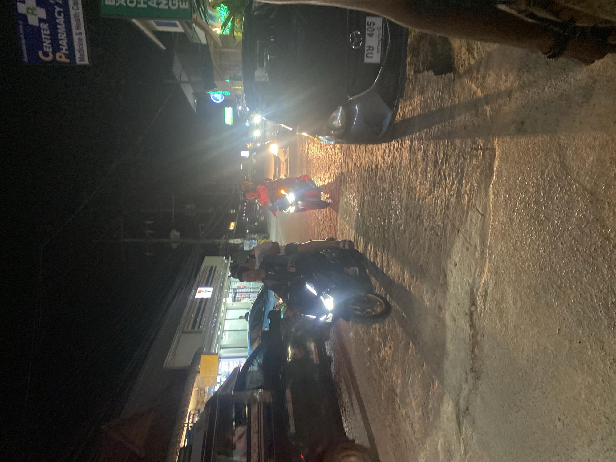 The image depicts a street scene at night, likely in a tropical or subtropical region. The ground appears wet, suggesting recent rain or high humidity. Several vehicles are present, including cars and a motorcycle.
In the foreground, a person is riding a motorcycle. The motorcycle's headlight is on, illuminating the wet road. Another motorcycle with a rider is visible further down the street, its headlight also illuminated. A dark-colored car is parked or stopped on the right side of the road, its license plate partially visible with Thai characters and the number "405". The car has a Mazda emblem.
In the background, there are several buildings with illuminated signs. One sign reads "CENTER PHARMACY" with "Medicine & Health Care" below it. Another sign appears to say "EXCHANGE" in green text. A store with a "7-ELEVEN" logo is also visible.
The overall impression is of a busy street at night, with activity from vehicles and commercial establishments. The lighting suggests it is dark outside, and the wetness of the road adds to the atmosphere. The presence of Thai license plates and store names indicates a possible location in Thailand.