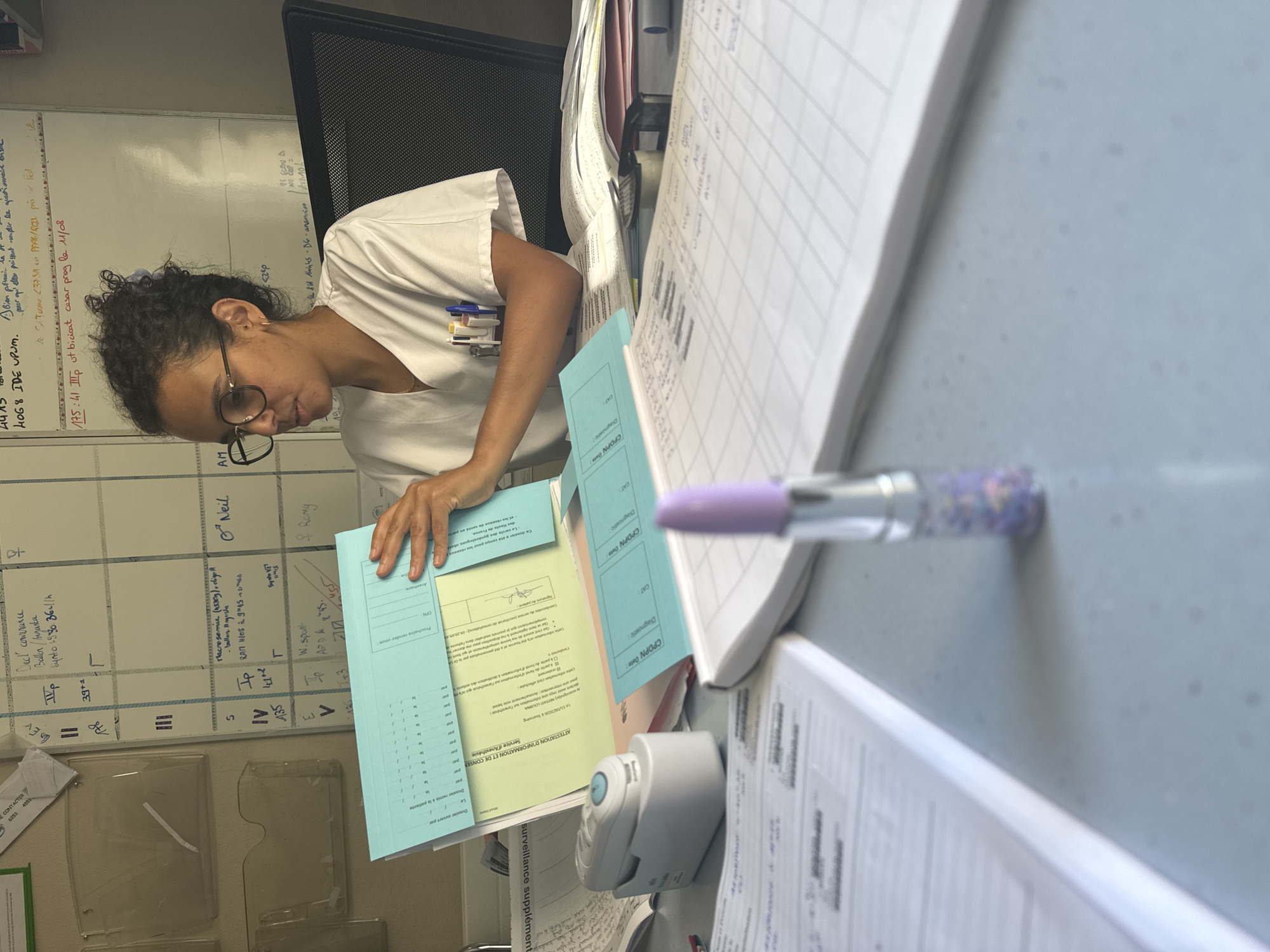 A young woman with curly dark hair and glasses, wearing a white medical uniform, sits at a desk in what appears to be an office or medical facility. She is looking to her right and holding a stack of blue and yellow documents. On the desk in front of her are several other papers, a stapler, and a tube of lipstick with a purple cap. Behind her, a whiteboard with handwritten notes is visible. The overall impression is of a workspace in a medical setting, possibly in Tourcoing, France, given the handwritten French text on the whiteboard and documents.