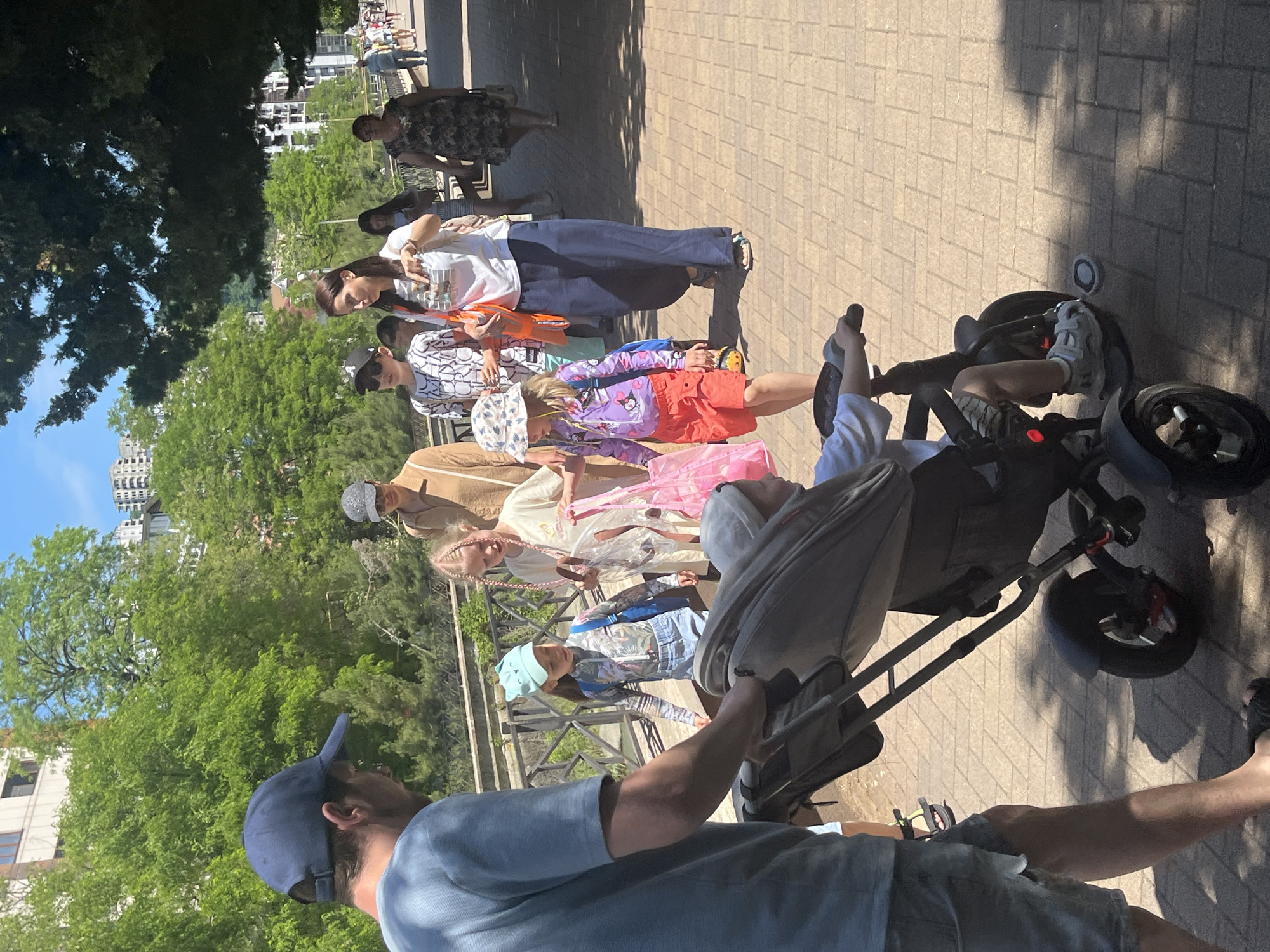 The image captures a group of people, including several adults and children, enjoying a sunny day outdoors in what appears to be a park or promenade in Sochi, Russia. The perspective is from behind a man in a blue cap and t-shirt, who is pushing a dark-colored stroller or child's push-trike with a bag attached. A child is seated in the stroller, with their legs visible.
In front of the stroller, a line of people is walking along a paved pathway made of light-colored interlocking pavers. This group includes a young girl in a patterned beanie and blue jacket, and another child in a purple cartoon-print jacket and red shorts. Several adults are also present, dressed in casual attire such as t-shirts, long pants, and skirts/dresses. One woman wears a white t-shirt with graphics and holds an orange bag. Another woman in a dark patterned dress is visible further down the path.
The setting is characterized by abundant green foliage and trees lining the left side of the path, with buildings visible in the background against a clear blue sky. Bright sunlight casts distinct shadows on the ground, indicating a daytime scene, likely late morning or afternoon. The overall atmosphere appears relaxed and casual, suggesting a leisurely outing. No specific readable text is clearly visible, though there are patterns and graphics on some clothing items.