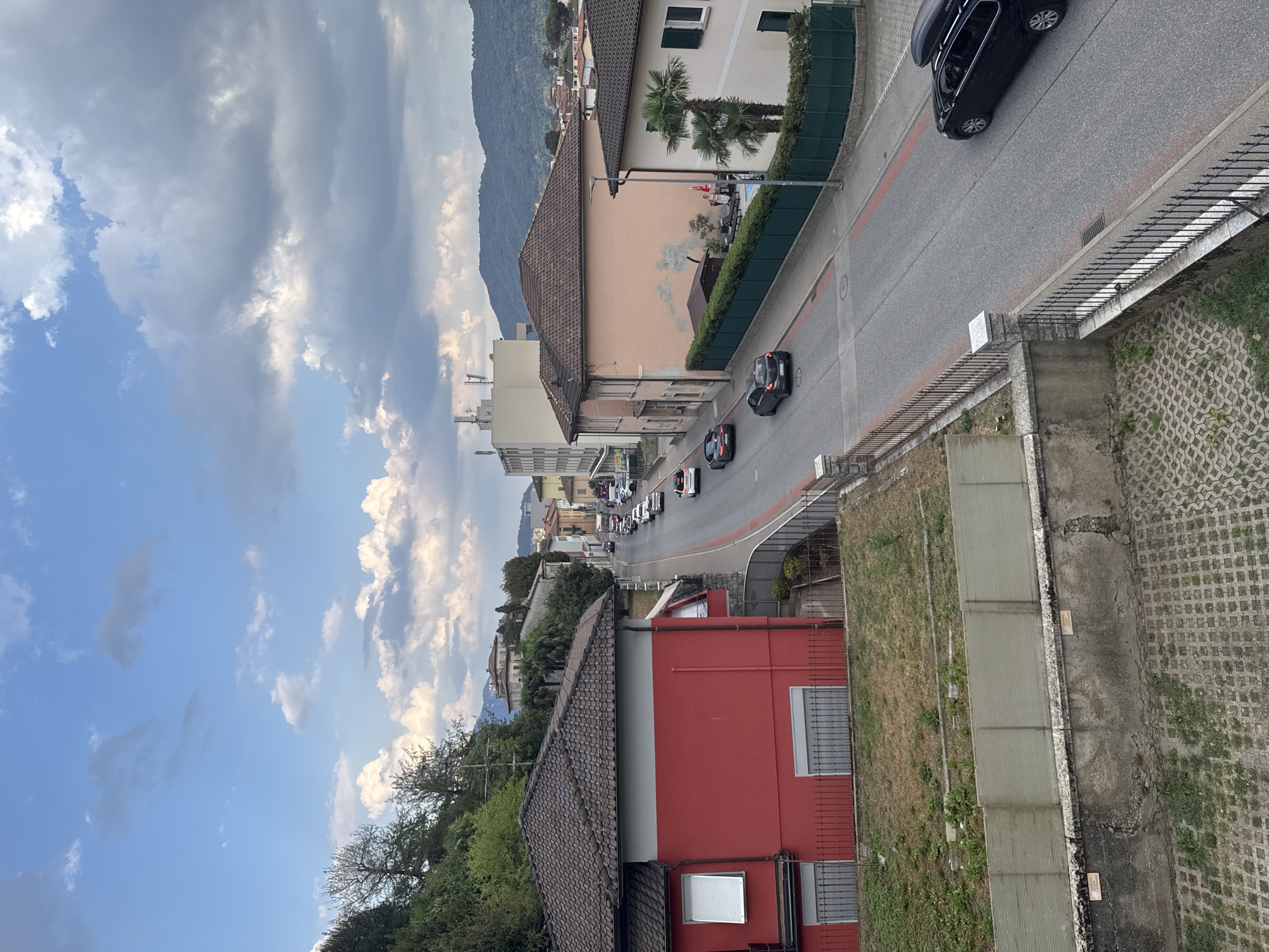 The image captures a street scene in Balerna, Switzerland, under a partly cloudy sky. A paved road runs through the center, bordered by sidewalks and a green fence on the right. Several cars are visible on the road, including a black SUV with a roof box parked on the right side. Buildings line the street, with a prominent red house on the left and a pale pink building with palm trees in front on the right. The foreground shows a concrete wall with a decorative patterned surface and some overgrown vegetation. In the distance, hills rise under a sky filled with dynamic cloud formations.