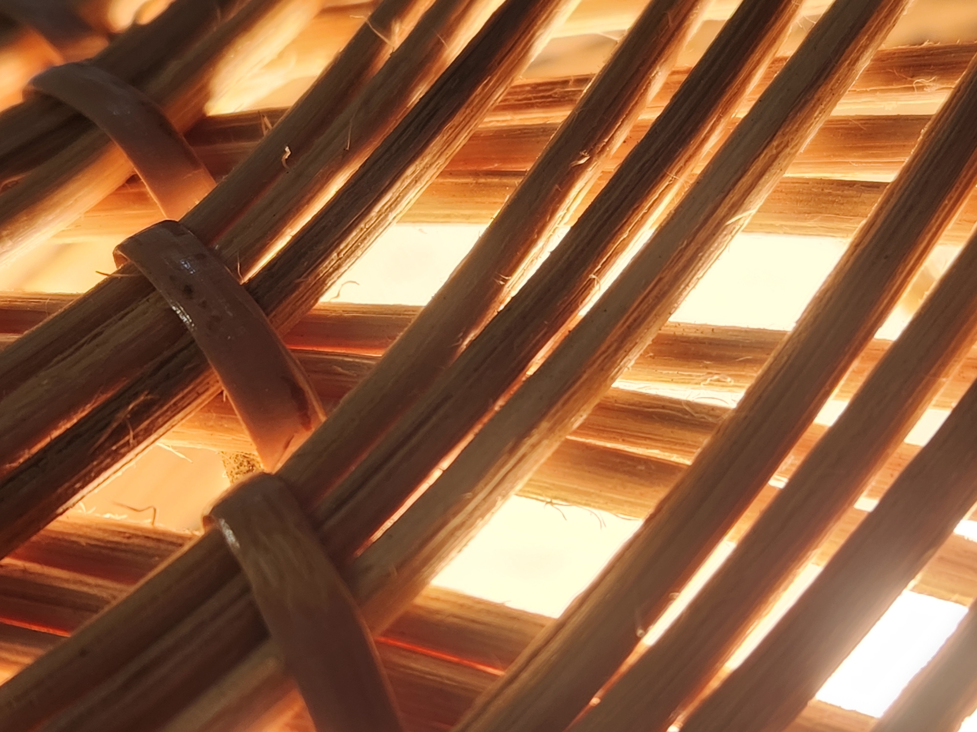 This is a close-up, abstract photograph of woven bamboo material illuminated from behind. The bamboo stalks are thin and appear to be naturally colored, with variations in tone from light tan to darker brown. The weaving pattern creates diagonal lines and openings through which a bright, warm light source is visible. The lighting casts shadows and highlights on the textured surface of the bamboo, creating a sense of depth and warmth. There are no people or specific environmental details that would indicate a location within Maitland, United States of America. The image is purely focused on the texture and pattern of the woven bamboo.