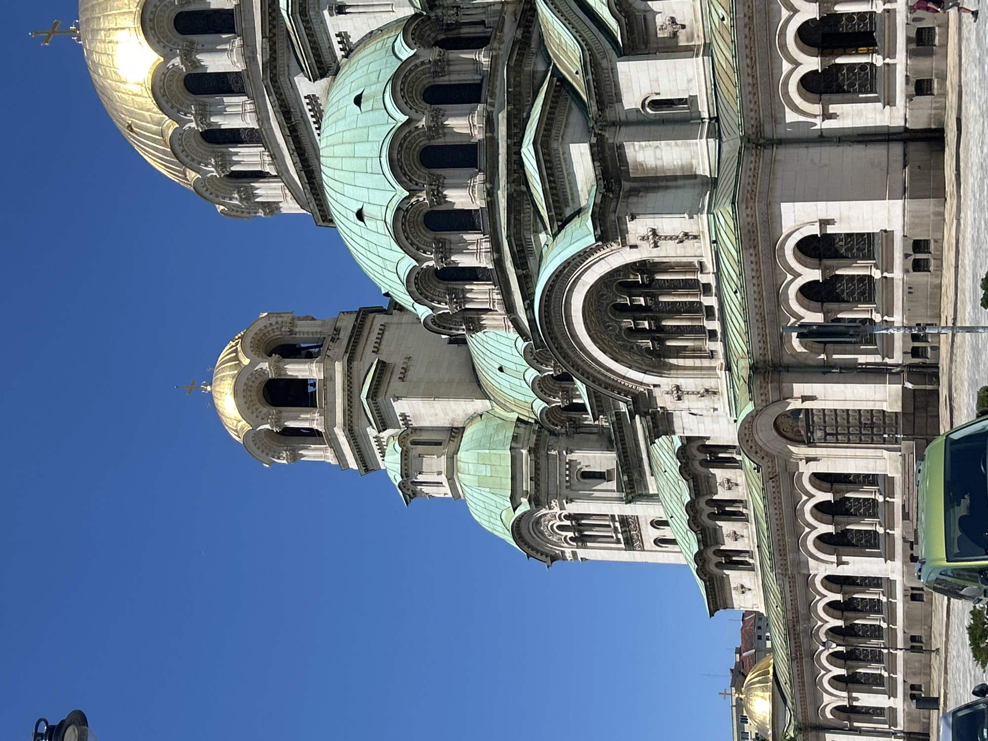An exterior, eye-level, vertical, medium shot captures the Saint Alexander Nevsky Cathedral in Sofia, Bulgaria, under a clear blue sky. The grand cathedral's iconic golden and green domes and intricate stone architecture dominate the frame. A street lamp is partially visible in the upper left corner, and two cars, one green and one silver, are parked in the lower portion of the image. The ground appears to be paved, and a few people are walking on the sidewalk. The sunlight casts shadows on the building, highlighting its detailed carvings and arched windows.