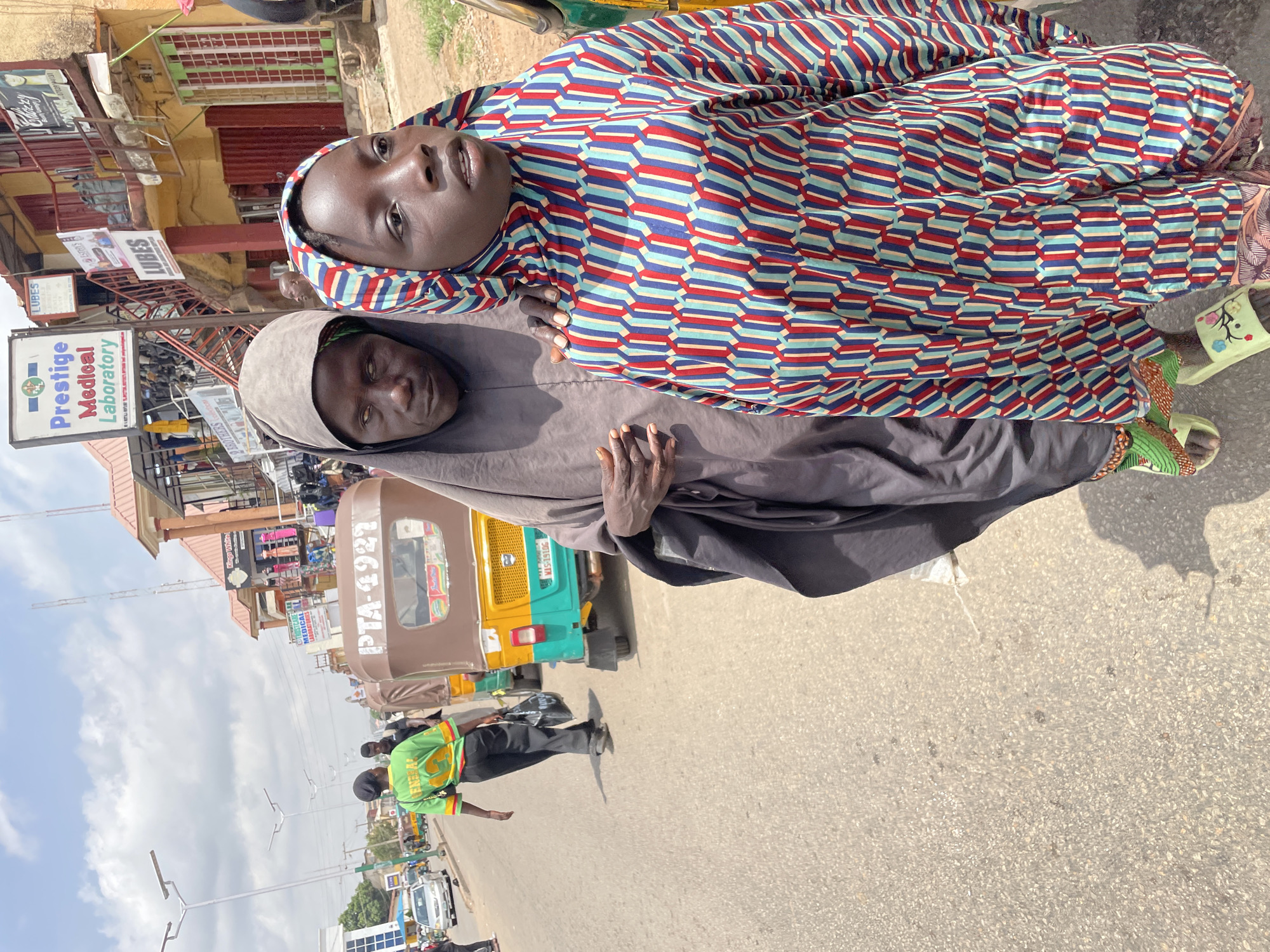 Two women are standing on a street in what appears to be Jos, Nigeria. One woman is wearing a colorful patterned dress and the other is wearing a gray hijab. They are positioned in front of a yellow-and-green "Keke Napep" taxi with license plate "PTA 4927" and "MIS-1910G." A man wearing a green and yellow t-shirt walks on the street beside them. Behind them is a building with a sign that reads "Prestige Medical Laboratory" and a partially visible sign that reads "Lubes." Other buildings and signs are visible in the background, suggesting a commercial area. The sky is partly cloudy.