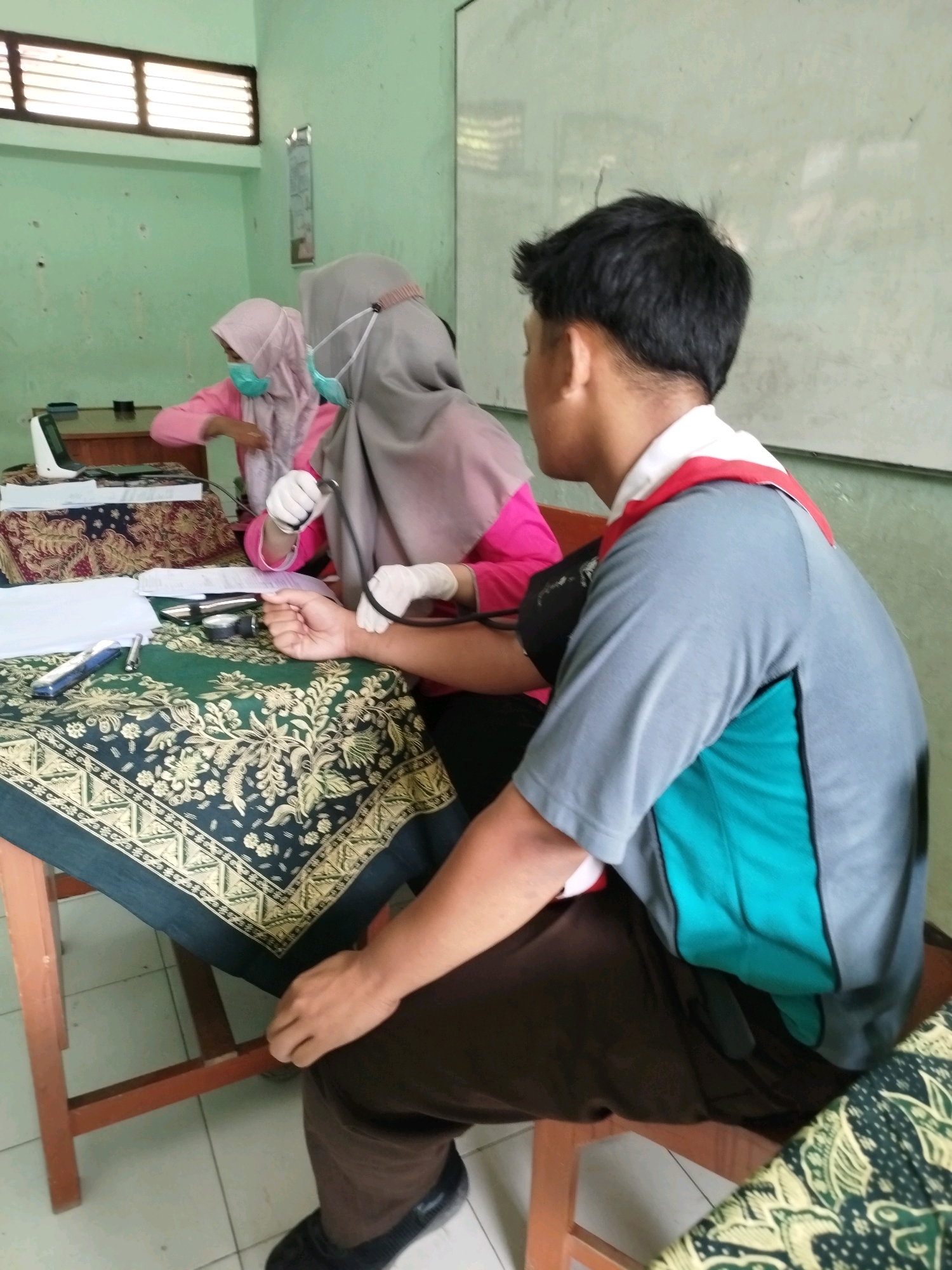In a room with light green walls and windows with horizontal slats, two young individuals are interacting at a wooden table covered with batik fabric. On the left, a person wearing a pink hijab and a light blue mask is operating a digital blood pressure monitor. In front of them, a patient, a young male wearing a grey polo shirt with a red collar and dark brown pants, has his arm extended to have his blood pressure taken. The blood pressure cuff is visible on his arm. To the right of the monitor, a whiteboard is partially visible on the wall. The scene appears to be a health check or screening in a school or community setting in Karangsambung, Indonesia.