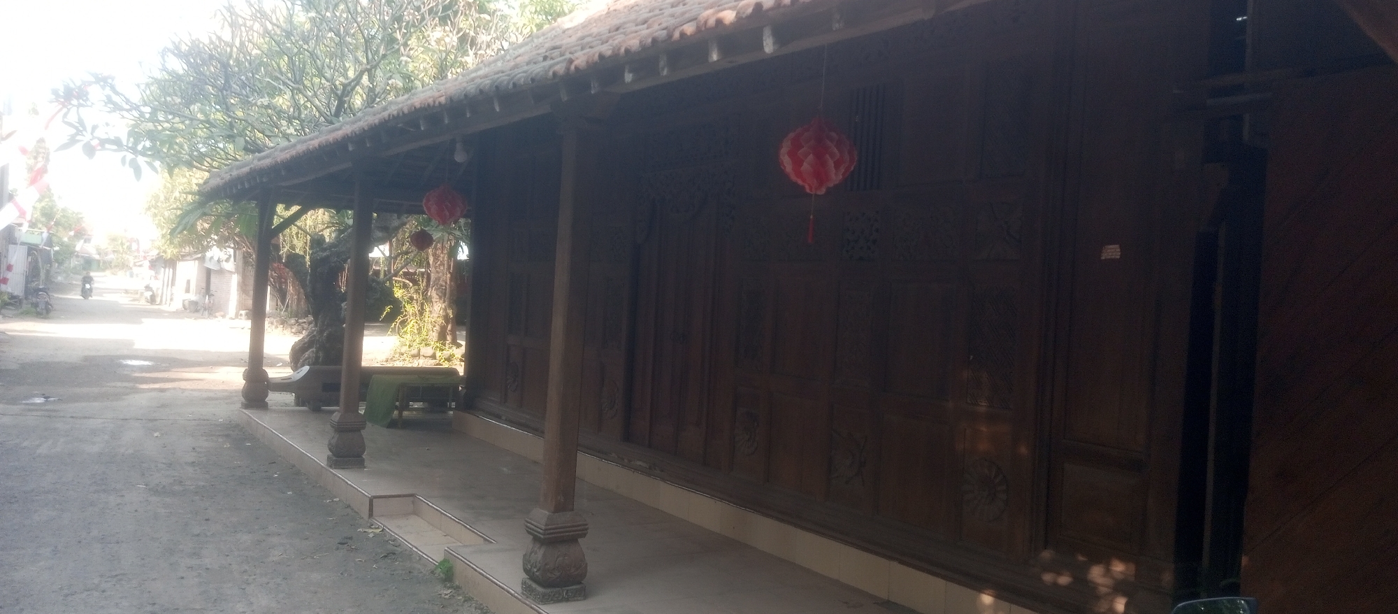 The image depicts the exterior of a traditional Javanese wooden house in Kediri City, Indonesia. The house features intricate carvings on its wooden walls and a tiled roof supported by wooden pillars. A covered veranda runs along the front of the house. Two red, hexagonal lanterns are hanging from the eaves, one in the foreground and another further back. In the background, a dirt road is visible with a few buildings and trees lining its sides. A motorcycle is parked on the left side of the road, and there are some plants and a bench on the veranda. The lighting suggests it is daytime, with sunlight casting shadows on the ground and the house.