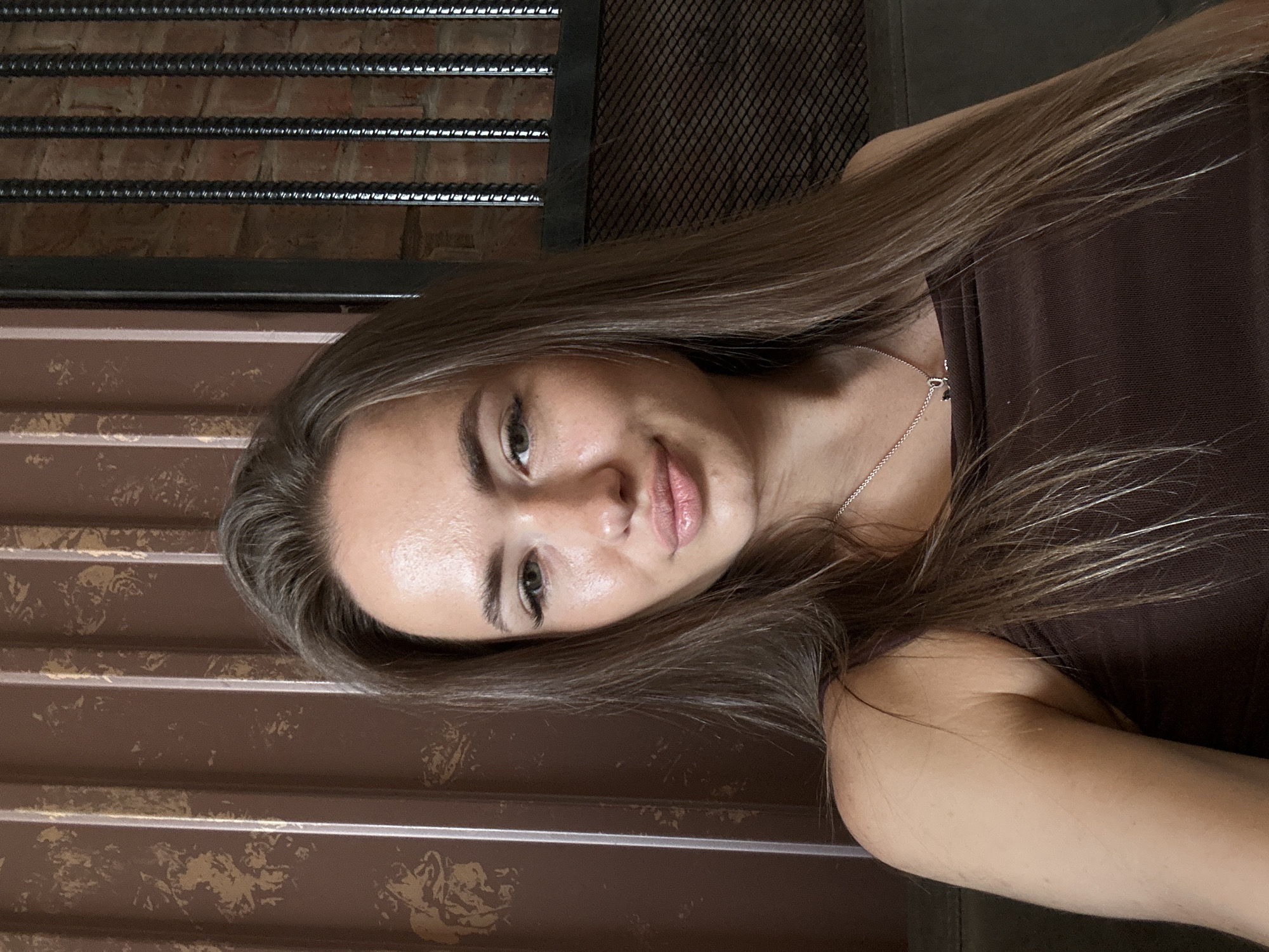 A young woman with long, straight, brown hair and fair skin is taking a selfie. She is wearing a brown tank top and a silver necklace with a small pendant. She has light green eyes and is looking directly at the camera with a slight smile. Her makeup is minimal, with emphasis on her eyelashes and lips. The background consists of a textured brown wall with vertical lines and a dark metal grate in front of a brick wall. The lighting is soft and natural, highlighting her facial features. The image appears to be taken indoors.