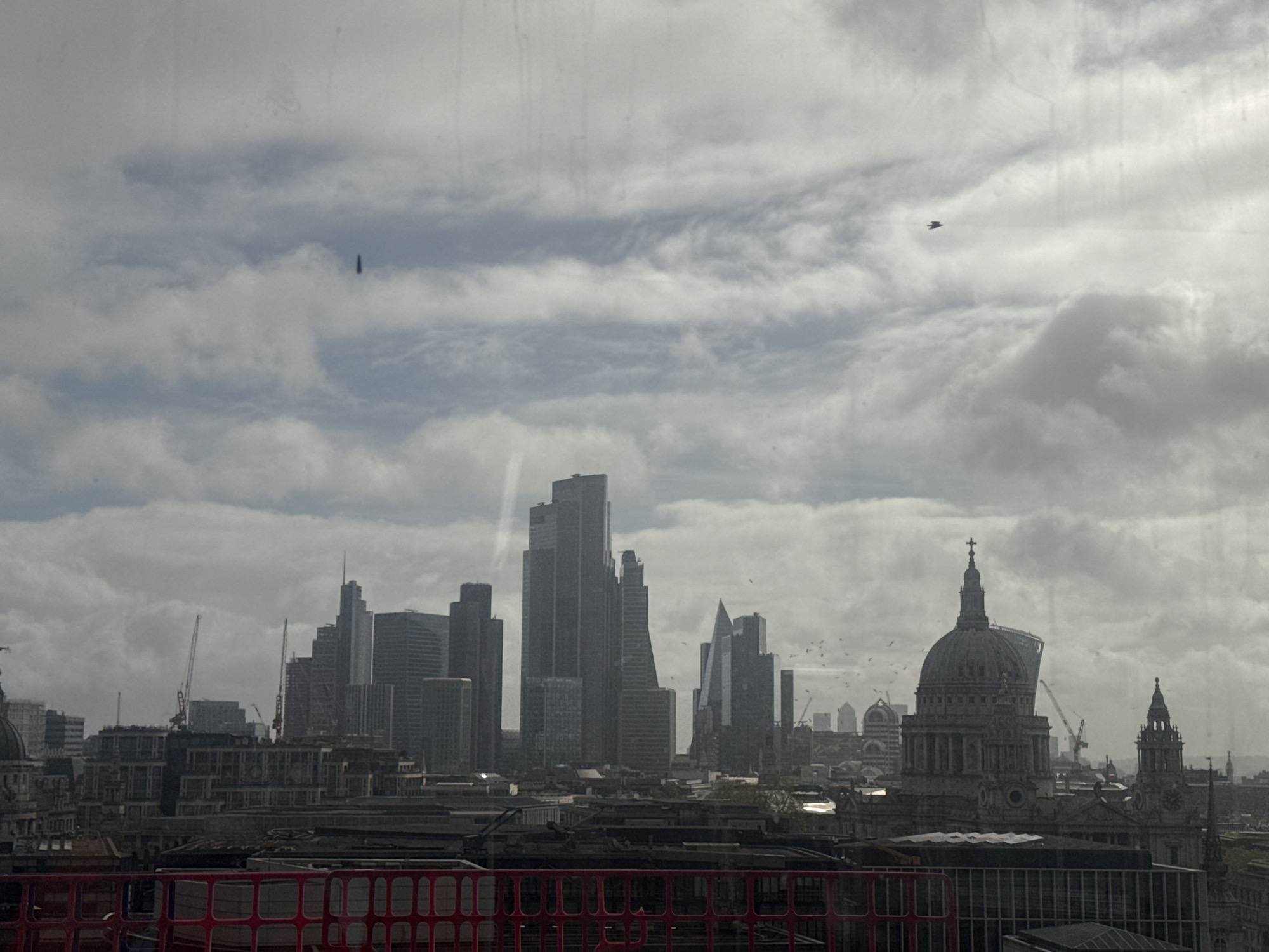 The image shows a cityscape in London, United Kingdom, under an overcast sky with heavy clouds. Prominent landmarks include the skyscrapers of the City of London financial district and St. Paul's Cathedral. Several construction cranes are visible among the buildings. A small, dark object, possibly a bird or aircraft, is in the sky. A red metal railing is in the foreground. The scene appears to be viewed from an elevated position.